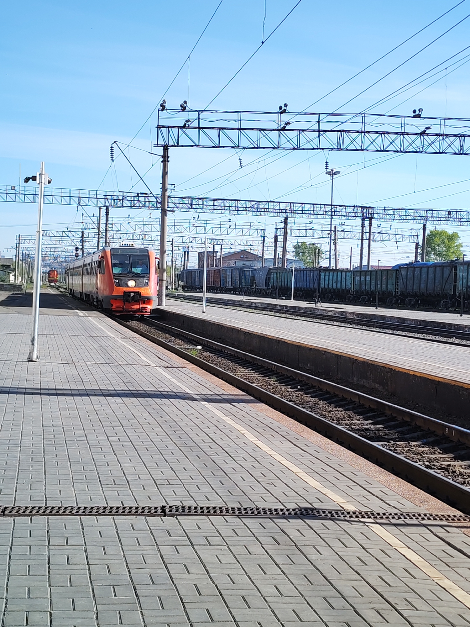 The image shows a train station in Izhevsk, Russia. An orange passenger train is arriving at the platform on the left. To the right, a train of cargo cars is parked on a track adjacent to the passenger platform. An overhead electrical grid supports the trains. The platform is made of grey rectangular paving stones and runs parallel to the tracks. A light pole stands at the edge of the platform, further left. The sky is blue.