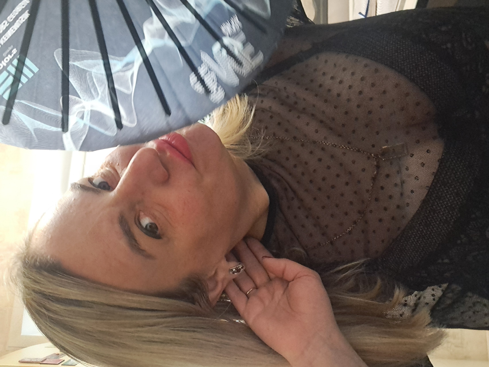 A woman with light blonde hair is seen in a close-up, tilted view. Her left hand is near her neck, displaying a ring on her finger. She wears a sheer, dotted top over a darker garment, with a thin gold chain necklace visible. Above her face is a partly obscured fan with black spokes and a light blue fabric displaying white designs and lettering including "...2 ENERGO KEMEKMAT..." and "DIO-2...". The setting appears to be indoors, and there's no explicit indication that this photo was taken in Elektrostal, Russia.