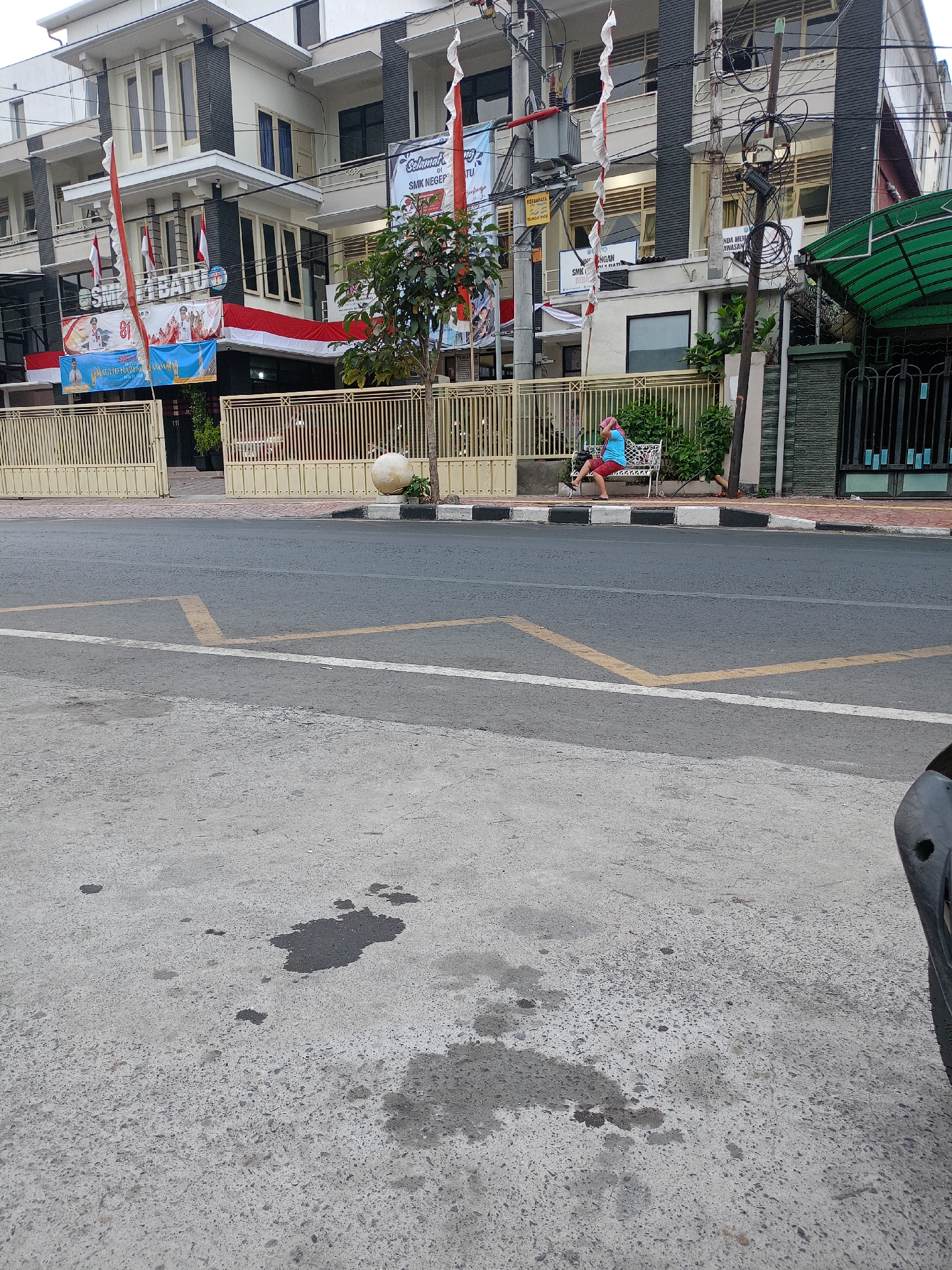 The image captures a scene in front of SMK Negeri 1 Batu, a vocational high school located in Batu City, Indonesia. In the foreground, a section of asphalt road is visible, marked with faded yellow lines and a white stripe. Some dark, irregular stains are present on the concrete pavement to the left of the road. Behind the road, a light-colored fence with vertical bars separates the sidewalk from the school building. A person is seated on a white bench to the left of the fence, facing away from the camera. This individual is wearing a light blue shirt and red shorts. The school building, a multi-story structure with a combination of white and dark grey facades, stands behind the fence. Several Indonesian flags and banners are displayed prominently on the building's exterior. Trees and electrical poles with numerous wires are also part of the environment. The overall impression is that of a street-level view of a public institution.