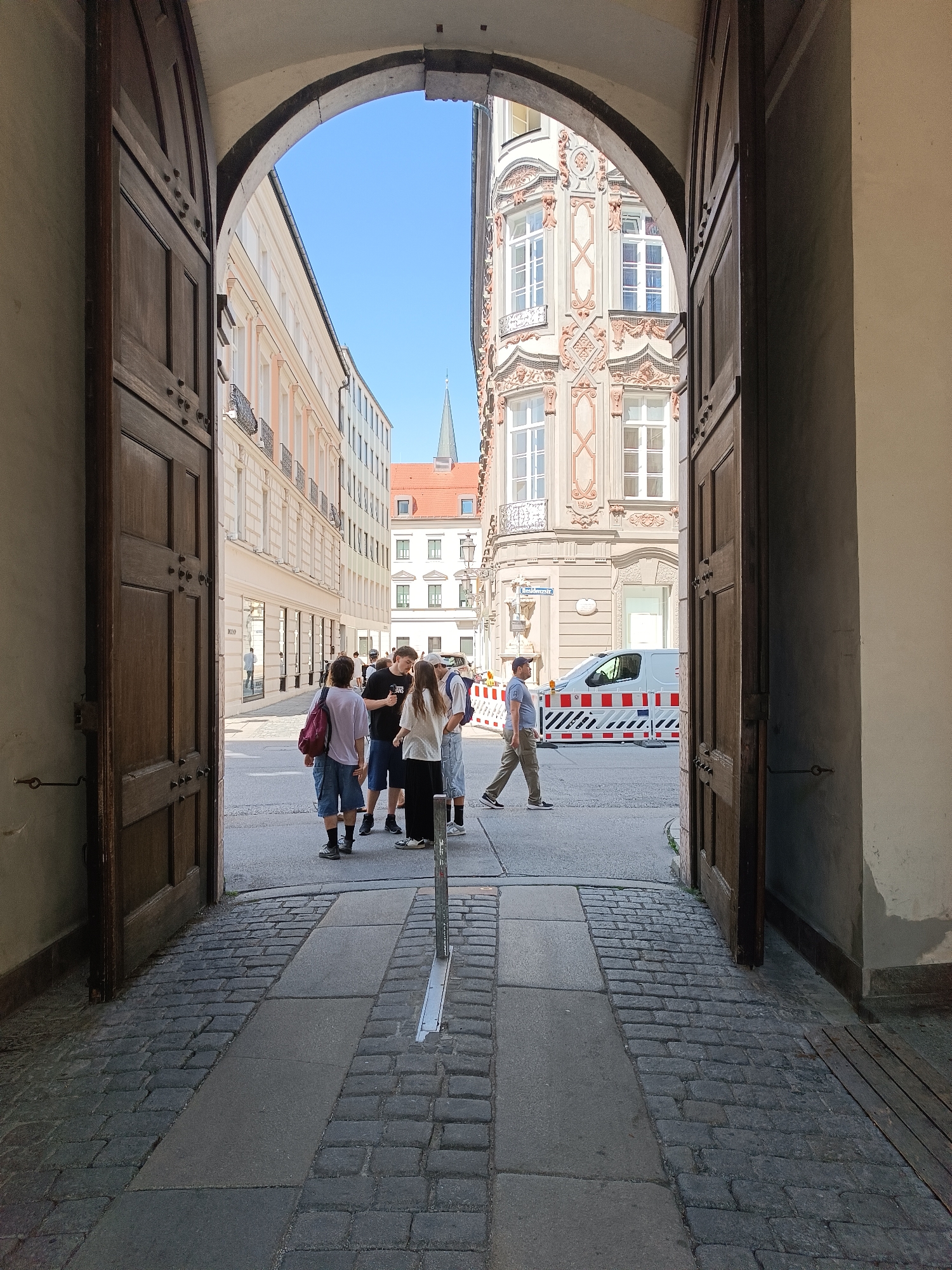 The image depicts an open archway of a building in Munich, Germany, looking out onto a city street. The archway is framed by large, dark wooden doors.
In the foreground, the ground is paved with cobblestones and a central path of large grey stone slabs.
Through the archway, on the street, several people are visible. On the left, a group of four individuals are standing and conversing. On the right, one person is walking away from the camera, and further down the street, another person is walking towards the camera. A white van with red and white barricades is parked on the right side of the street.
The buildings lining the street are multi-story, with architectural details characteristic of European city centers. The sky above is clear and blue. The scene suggests a sunny day in an urban environment.