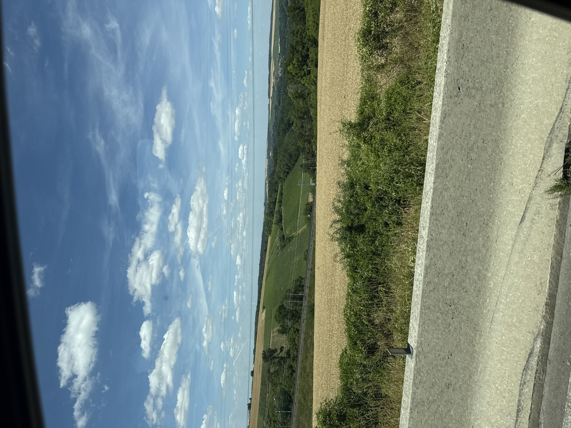 The image captures a landscape scene, likely from a moving vehicle, with a concrete barrier in the foreground. Beyond the barrier, a field of golden wheat stretches across the mid-ground. Further in the distance, rolling hills are covered in lush green vegetation, possibly vineyards or other crops, interspersed with trees. A railway track with overhead power lines runs horizontally through the middle distance, suggesting an active transportation route. The sky is a vibrant blue, dotted with fluffy white clouds. The overall impression is a pastoral, rural setting, possibly in the vicinity of Poilly, France, given the agricultural landscape and the presence of vineyards. There are no people or discernible human activity in the frame.