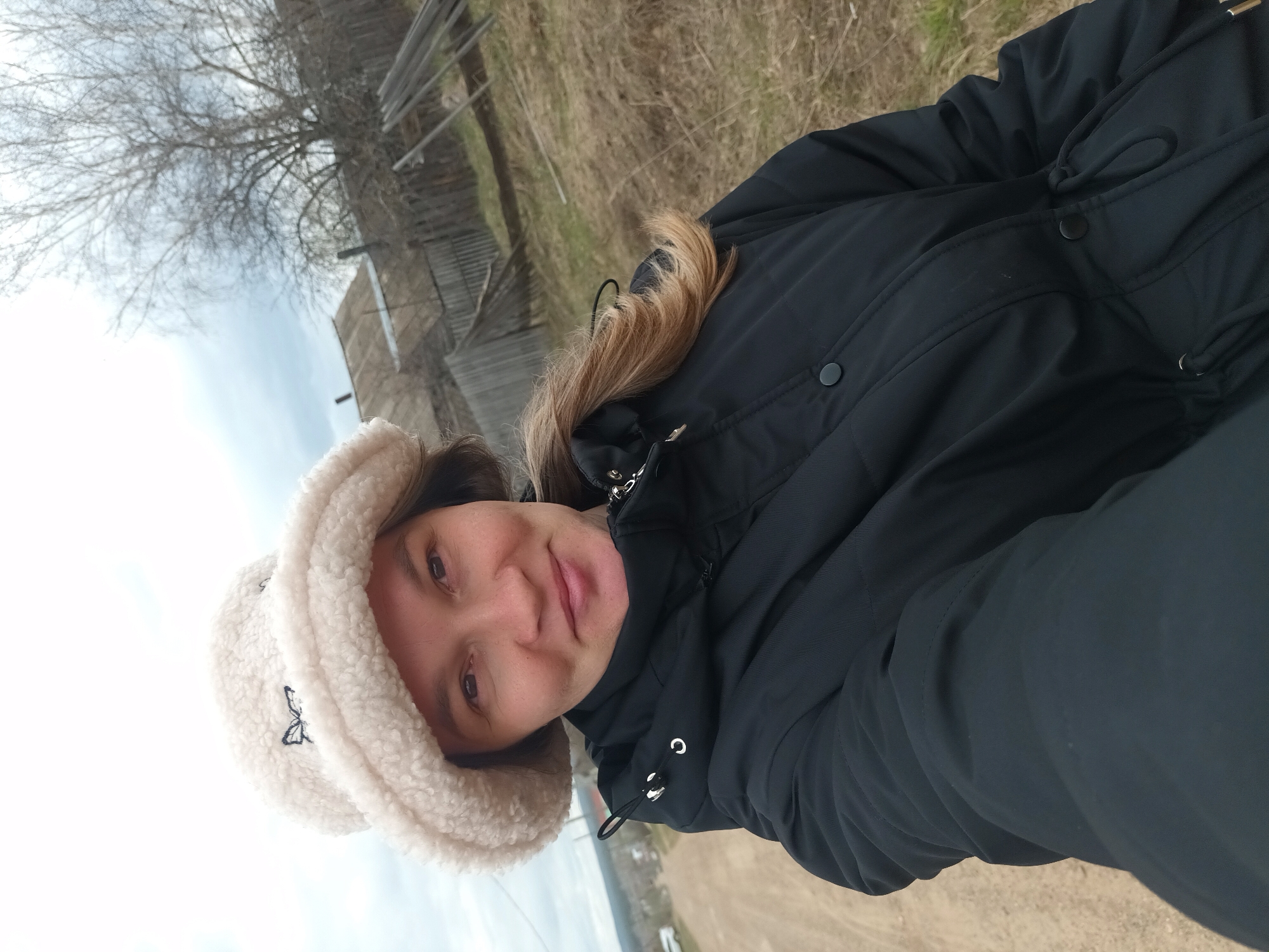 A woman is posing for a selfie outdoors, likely in the vicinity of Ольховка, Russia, as indicated by the presence of characteristic rural structures and landscape elements. The woman is wearing a black puffy winter coat and a white furry hat adorned with a dark-colored butterfly design. The background reveals a wintry rural scene with barren trees, a wooden fence structure, and hints of distant buildings. The sky is overcast, suggesting a cloudy day. The presence of dry grass and withered vegetation indicates a late autumn or early spring setting.