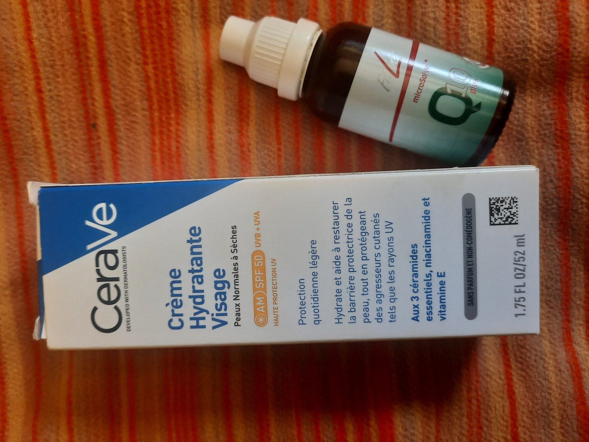 The image shows two cosmetic products placed on a textured, orange and red striped surface. On the left is a white and blue box with the brand name "CeraVe" prominently displayed. The box has French text indicating it is a "Crème Hydratante Visage" (Face Moisturizing Cream) with SPF 50. Below this, there is smaller text describing the product's benefits and ingredients, including "3 ceramides," "niacinamide," and "vitamin E." At the bottom of the box, it indicates the volume as "1.75 FL OZ/52 ml" and features a QR code. To the right of the box is a small, dark glass bottle with a white cap. The bottle has a label with a red stripe at the top and a green section featuring the text "Q10 plus" and "microSolve." The lighting in the image suggests it was taken indoors.