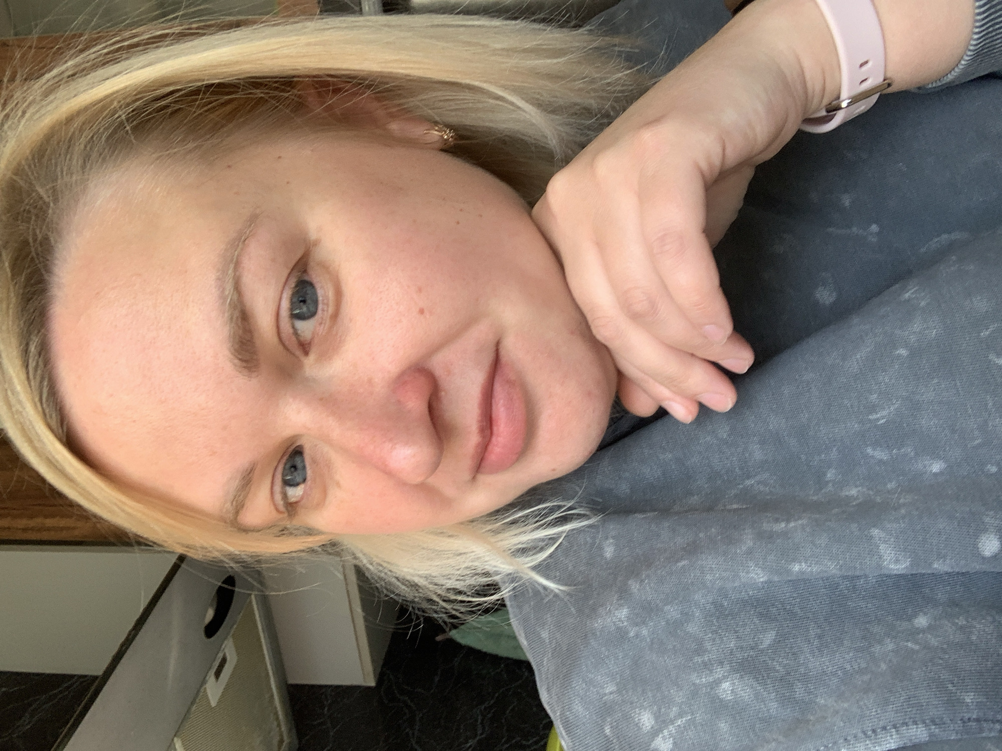 The image captures a casual, close-up self-portrait of a woman indoors, presumably in Chaikovsky, Russia. The main subject is a woman with blonde hair and clear blue eyes, looking directly at the camera. Her complexion is light, with a natural flush on her cheeks and nose, and some freckles. Her lips are slightly parted in a soft, pleasant smile, and she has light, natural eyebrows. She wears a small, intricate gold-colored earring in her right ear.
Her head is tilted to the right, resting on her left hand, which is visible. On her left wrist, she wears a light pink watch with a silver buckle. She is dressed in a grey long-sleeved sweatshirt or top, characterized by a textured, possibly acid-washed or distressed pattern.
The background is softly blurred but reveals an indoor setting. To the left, there's a light-colored surface, possibly a countertop or furniture, and a silver-toned electronic device lying flat. The floor appears dark with a subtle pattern. The lighting is soft and even, suggesting an indoor daytime setting. The woman conveys a relaxed and amiable emotion. There is no visible text in the image.