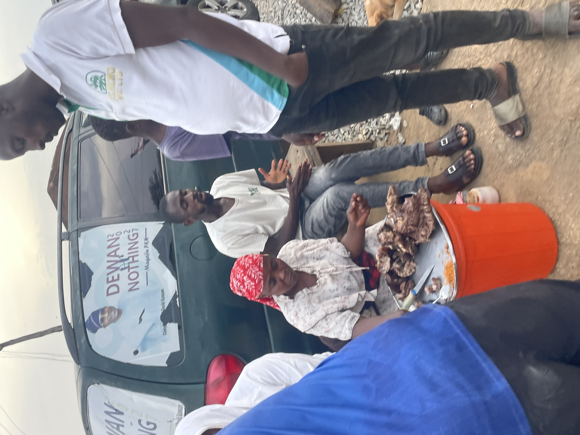 A street scene in Jos, Nigeria, shows a group of people gathered around a car. A woman wearing a red headscarf is sitting near an orange bucket, which contains pieces of meat. A man is sitting with his legs outstretched, gesturing with his hands. The back of a dark green car has a poster that says "DEWAN 2 DO NOTHING". Multiple people are standing or milling around the car. The ground appears to be dirt or gravel.