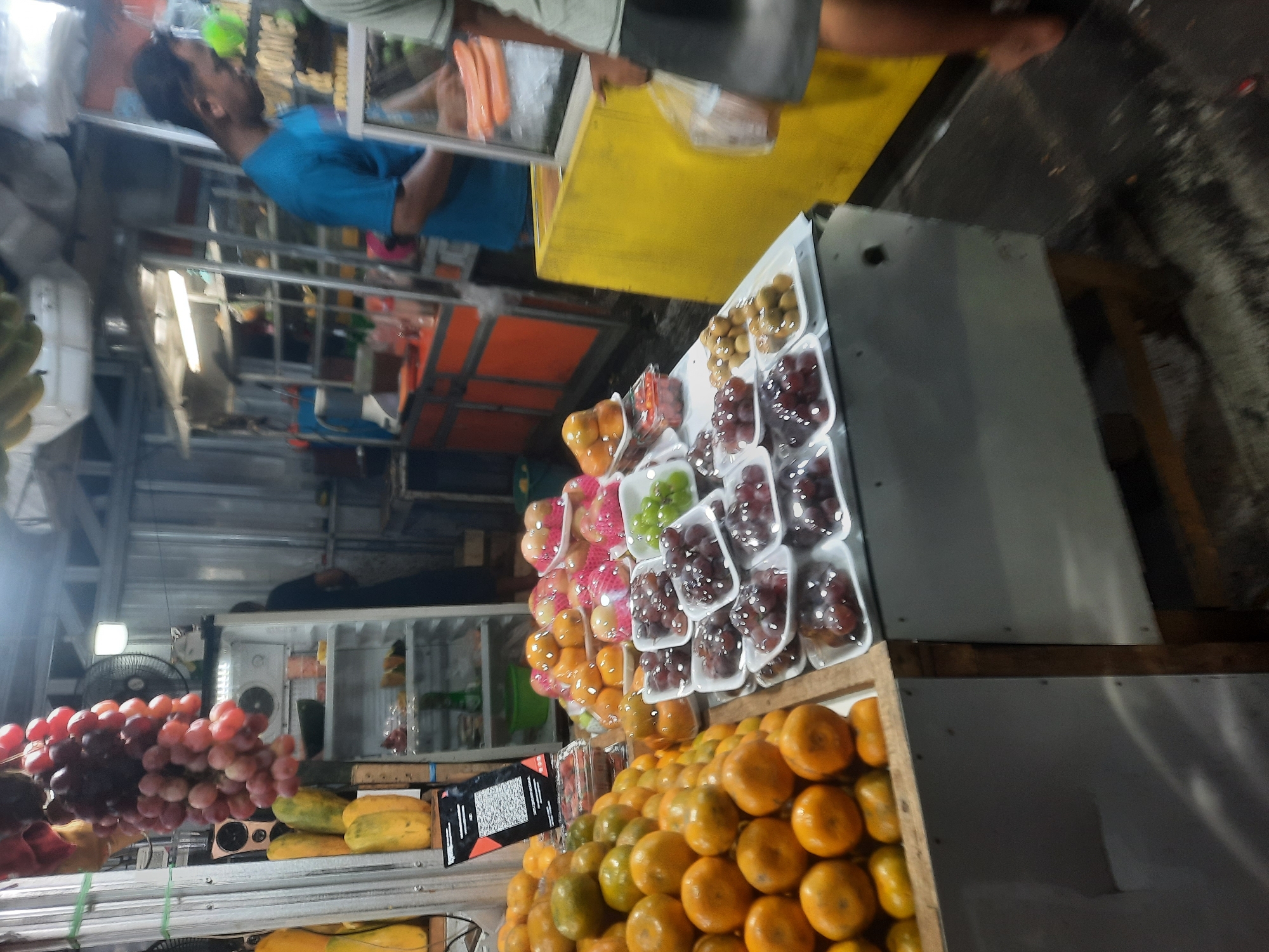 The image depicts a fruit stall at a market in North Jakarta, Indonesia. The stall is laden with various fruits, including oranges, grapes, and other produce displayed in plastic containers. In the background, a refrigerator and shelves filled with more items are visible, suggesting a bustling market environment. Two men are present; one in a blue t-shirt appears to be attending to customers, while another man, partially visible, is walking away. The lighting in the scene is artificial, contributing to the market's lively atmosphere.