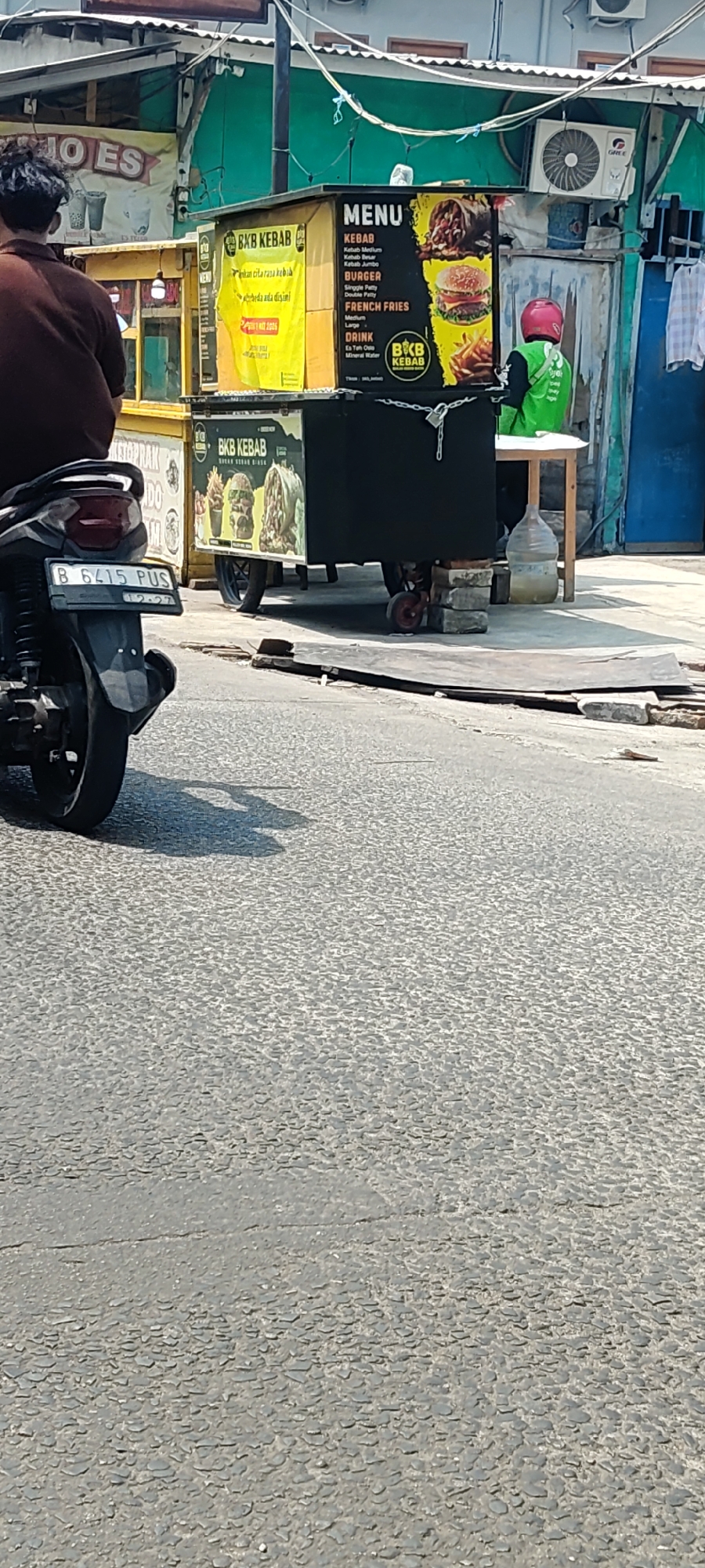 In Mekarmukti, Indonesia, a street scene unfolds with a parked motorcycle in the foreground, its license plate reading "B 6415 PUS". Behind it, a food cart, branded "BKB KEBAB," is prominently displayed. The cart features a colorful menu with illustrations of kebabs, burgers, and french fries. Adjacent to the cart, a person wearing a green uniform and a pink helmet is visible, suggesting a food delivery service. The environment appears to be an urban street with buildings and overhead wiring. The overall scene depicts a typical street-side food vendor operation in Indonesia.