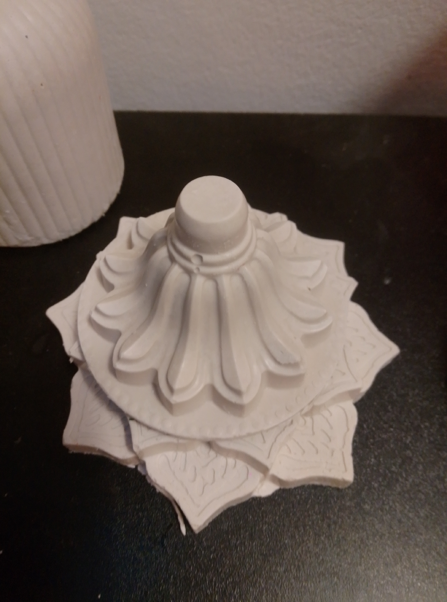 A white, ornate, plaster decorative element sits on a dark, textured surface. The element has a tiered, floral design with carved details. In the background, to the left, is a portion of a ribbed white object, possibly a vase or a pillar. The scene is indoors, with a light gray wall visible in the upper background. The photograph was taken in Wedel, Germany.
