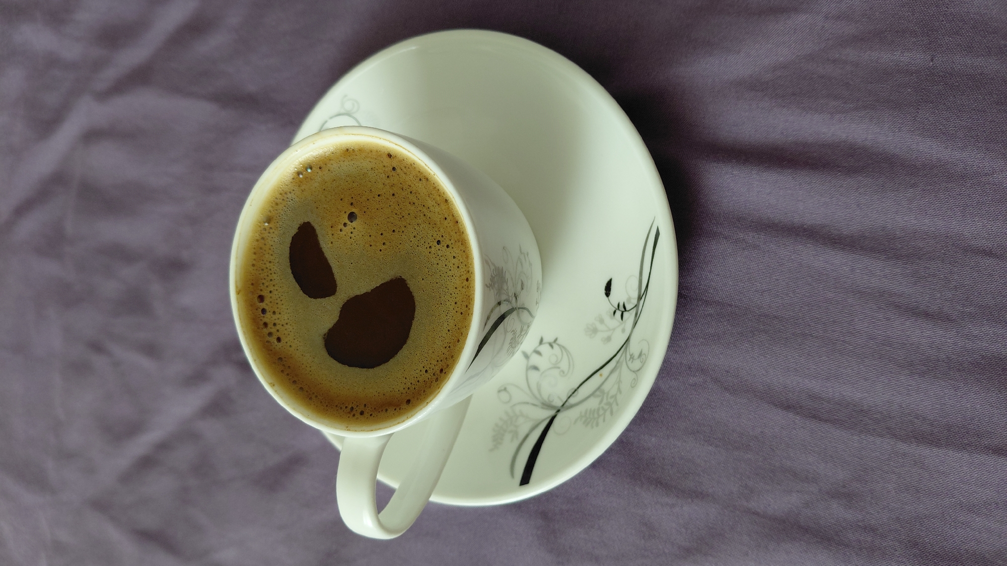 A white ceramic cup filled with dark coffee sits on a matching saucer with a delicate black and gray floral design. The coffee has a thick layer of foam, with two darker patches on the surface creating a subtle smiley face. The cup and saucer are placed on a textured, purple fabric surface. The image is a close-up shot, with the majority of the frame occupied by the coffee, cup, saucer, and fabric. The lighting is soft, casting gentle shadows. The scene appears to be indoors, possibly on a table or bed. There is no indication of the location being in the city of Abovyan, Armenia.