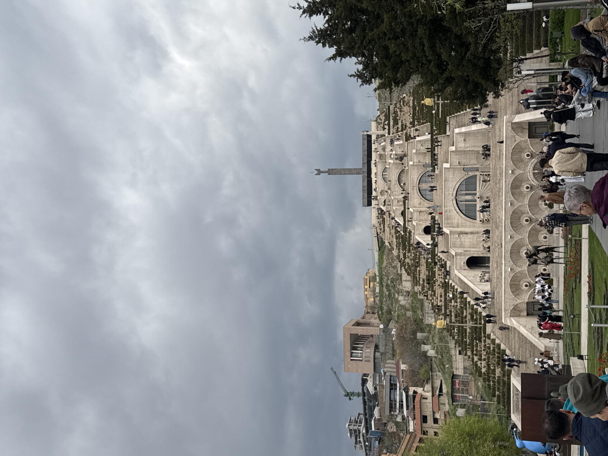 The image shows the Yerevan Cascade in Armenia under a cloudy sky. The Cascade is a massive, tiered, white stone staircase structure that ascends a hillside. Numerous people are seen walking up and down the stairs. Green spaces are visible along the sides of the staircase. Modern buildings are at the base of the Cascade, with a construction crane present. The sky is overcast, with dark clouds dominating the upper portion of the image. A statue stands prominently at the top of the Cascade. Groups of people are gathered at various levels.