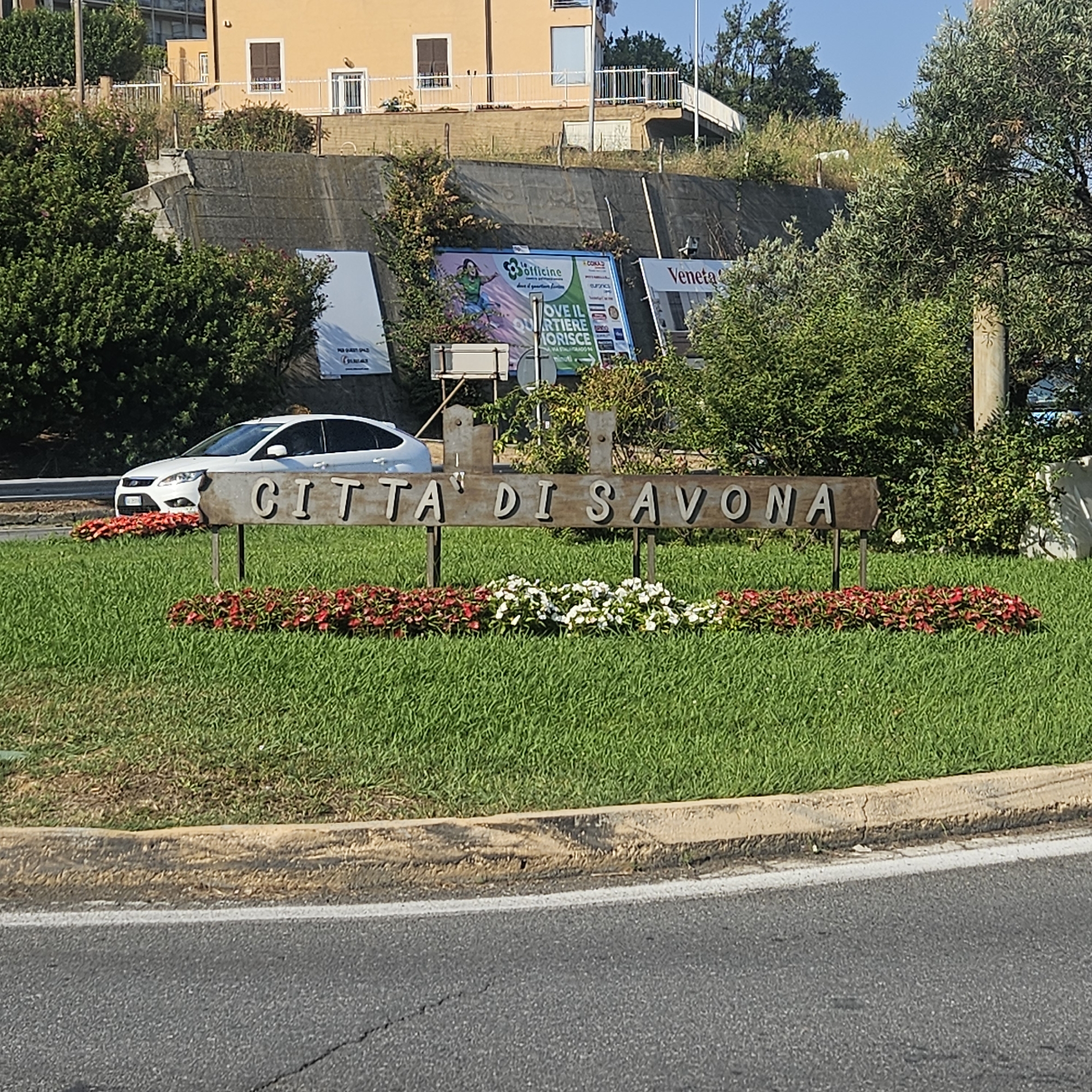 The image shows a street-level view of a sign that reads "CITTA DI SAVONA" in Italian, indicating the city of Savona, Italy. The sign is made of weathered wood with individual metal letters. It is mounted on two posts in a grassy area with red and white flowers planted in front. To the left of the sign, a white car is parked on the side of a road. In the background, there is a sloped retaining wall with advertisements and vegetation growing on it. Above the wall, a multi-story building with yellow walls and brown window frames is visible. The sky is clear and blue. The overall scene suggests a roadside entrance to the city, likely in an urban or suburban area.