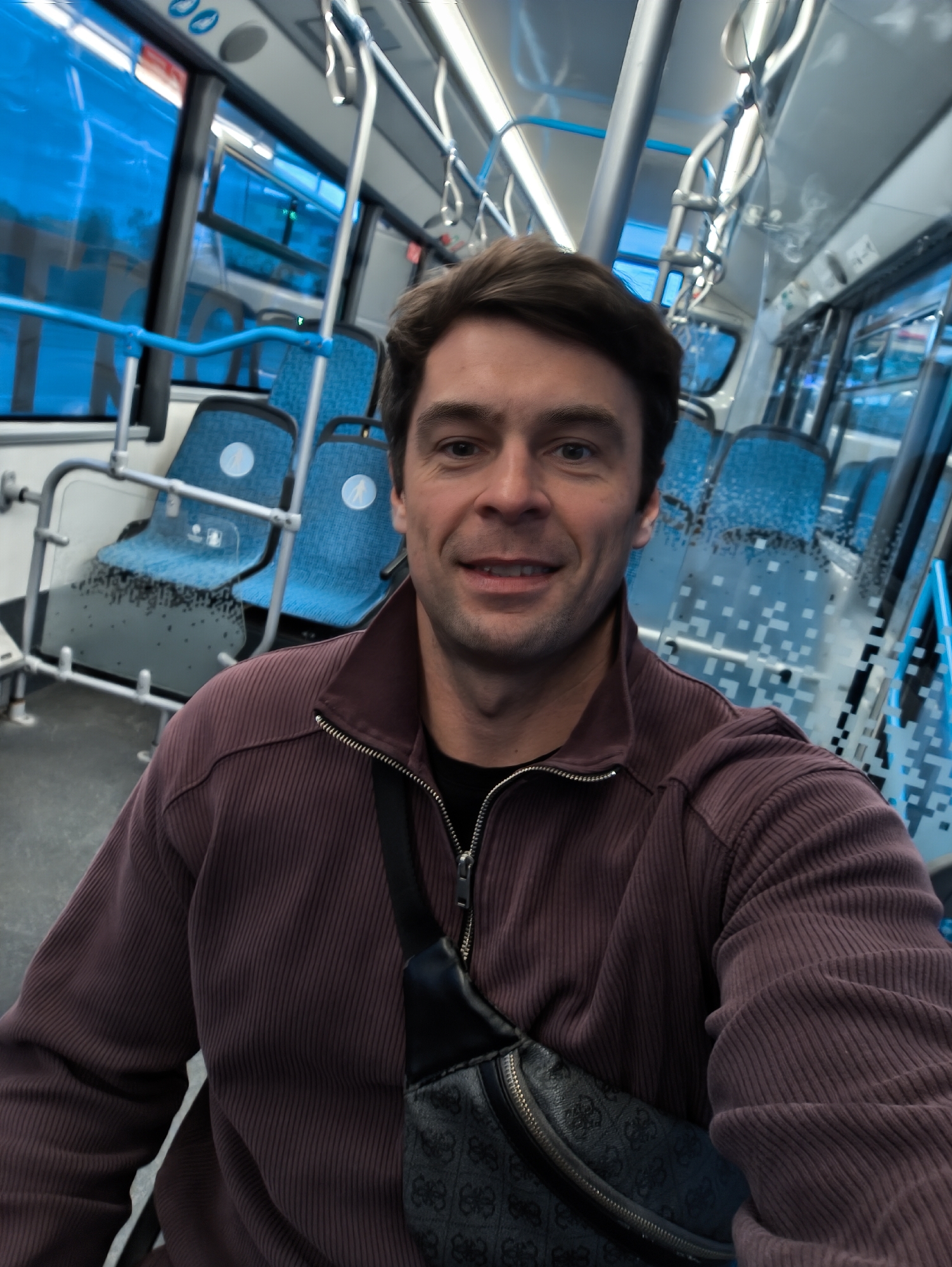 A man in a burgundy, vertically-ribbed jacket and a black patterned fanny pack is taking a selfie on public transportation. He is indoors, possibly on a bus or tram in Moscow, Russia. This is indicated by the blue interior, seating, and window design, which is typical of Moscow public transport. The man is facing the camera with a slight smile. Seating visible in the background. The scene is well-lit.