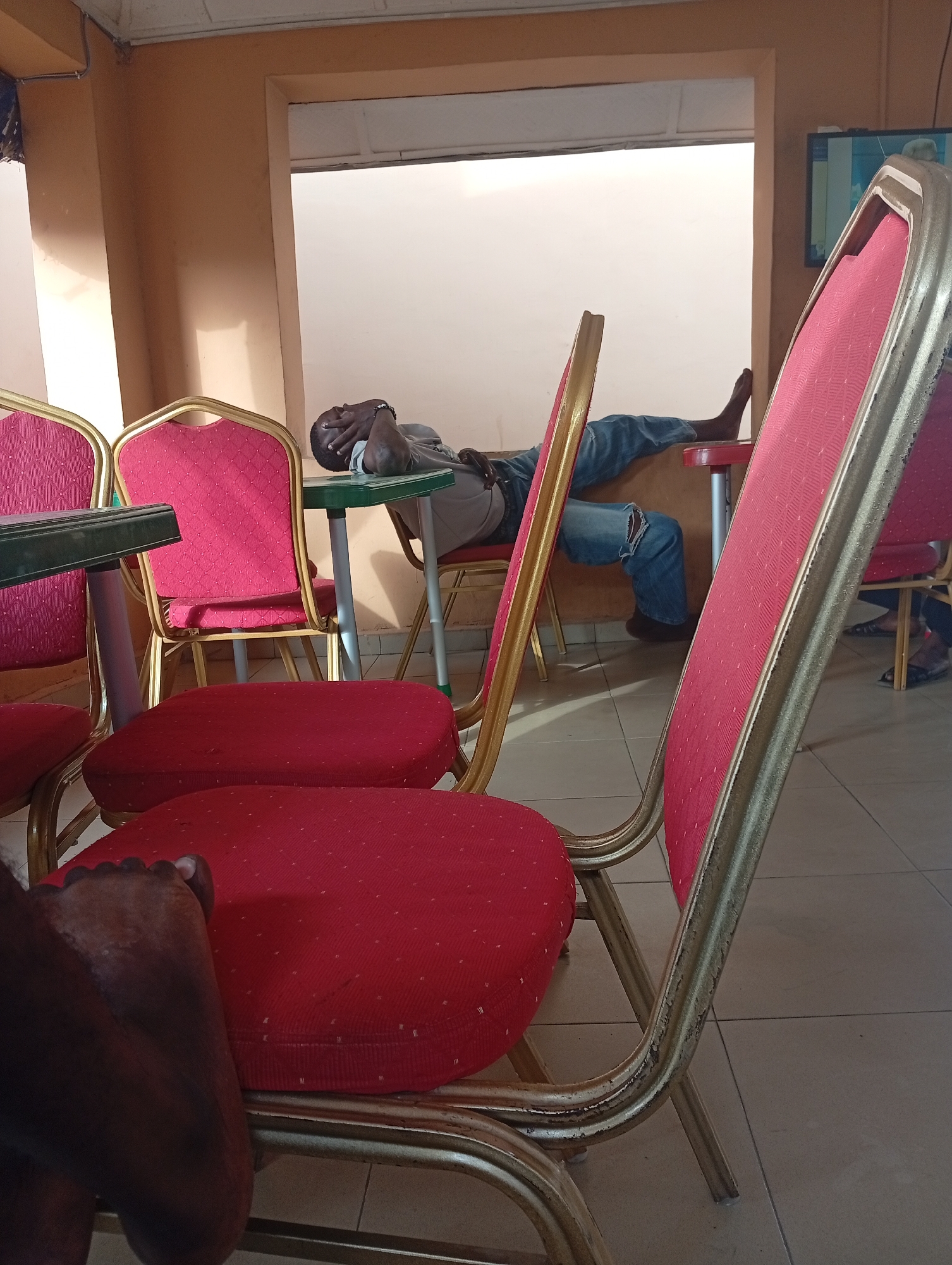 The photograph shows a room in Lokoja, Nigeria, furnished with multiple gold-framed chairs upholstered in red fabric. One man is sleeping, with his head on a small green table, legs extended onto another table. He wears jeans with a rip on the knee and a grey t-shirt. A television is partially visible on a wall. Another person's foot is visible in the foreground, near the base of one of the chairs. The floor is tiled. The room is bright, suggesting daylight.