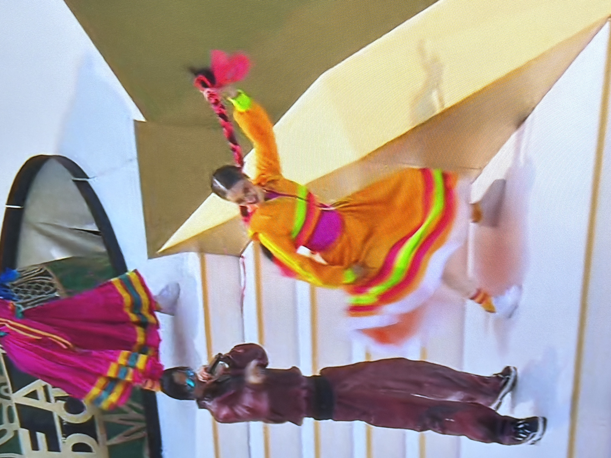 The image shows three people dressed in traditional Bulgarian folk costumes performing on a stage. The person in the foreground is wearing a brown, long-sleeved shirt and dark trousers, with black and white sneakers. They are looking down and appear to be holding something small in their hands. To the left, another person is wearing a vibrant pink, ankle-length skirt with yellow and blue stripes at the hem, along with a matching top. The skirt is full and pleated, suggesting it's part of a dance costume. In the center and background, a third person is captured mid-motion, wearing a bright orange dress with vibrant pink, lime green, and yellow trim. Their arms are outstretched, and they have a braided red hairpiece. The setting appears to be indoors, with white walls and a sloping ceiling. Shadows suggest artificial lighting. The overall atmosphere seems lively, indicative of a cultural performance or festival. No text is visible in the image.