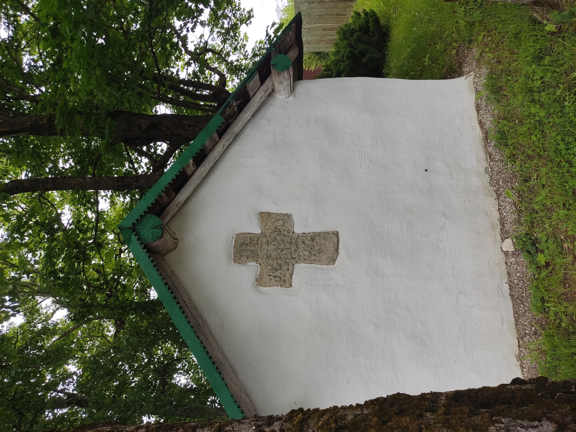 A small, white, building with a gabled roof featuring green trim and a cross embedded in the wall is partially obscured by a tree in Izborsk, Russia. The building is flanked by greenery and a wooden fence. The top of the tree and the sky are visible above the structure. The building appears to be in a natural setting.