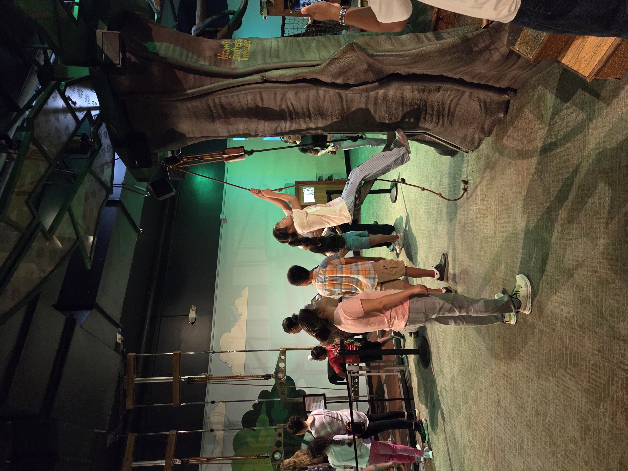 The image depicts a group of people, including children and adults, inside an indoor attraction, possibly an interactive museum or a play area. The setting appears to be themed around a treehouse or forest environment, with a large, faux tree trunk as a prominent element. A young girl is actively engaged in climbing or swinging from a rope attached to the tree, showing an upward motion. Several other children and adults are observing, some standing in a queue or line. The floor is carpeted with a textured pattern. The lighting is artificial and casts shadows. A sign with yellow lettering is visible on the tree trunk, reading "Horowitz Brothers Treehouse." In the background, there are hanging structures and what appears to be a painted mural of clouds and trees on a wall. The overall atmosphere suggests a recreational or educational activity for families.