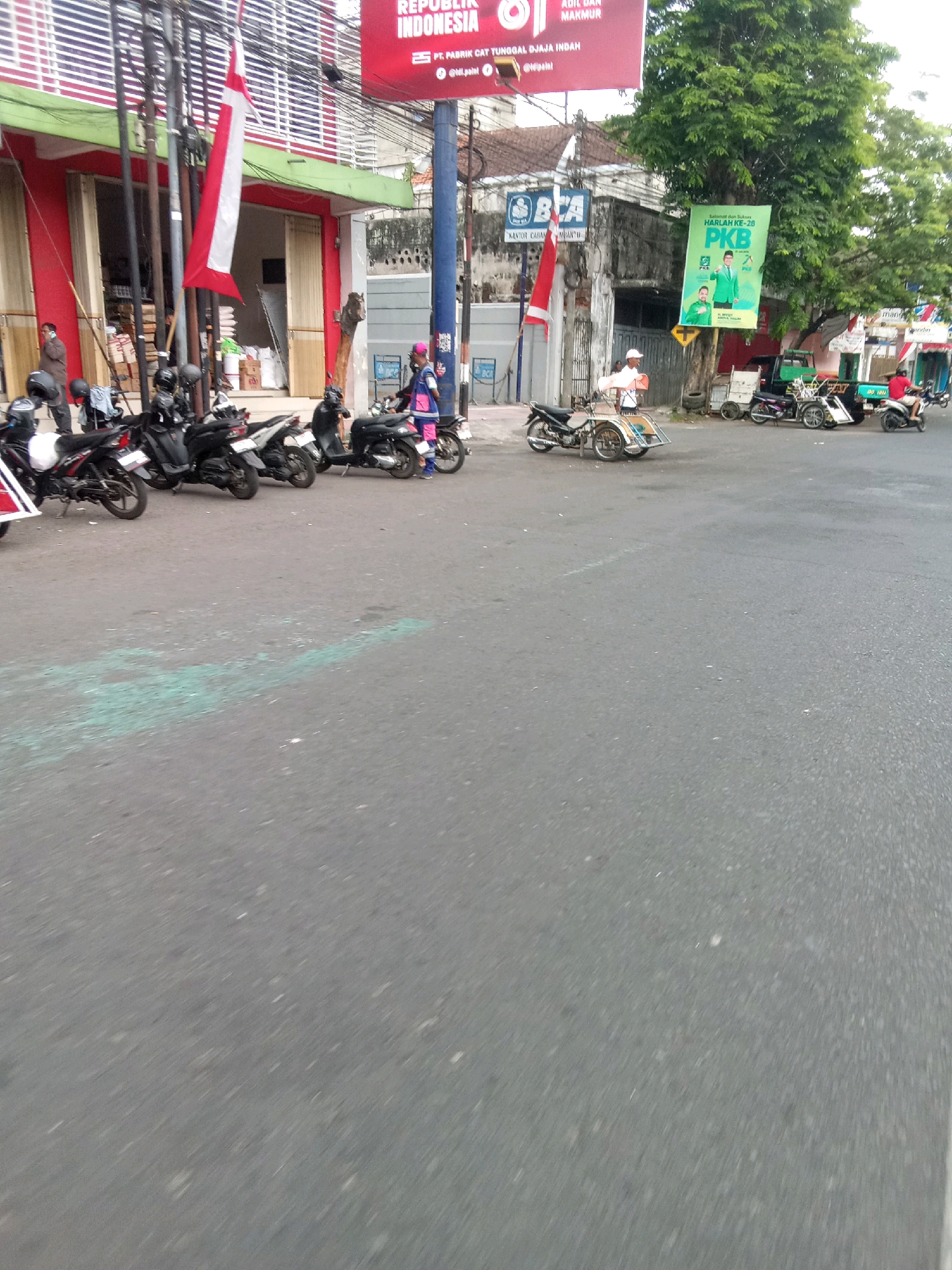 The image shows a street scene in Jember, Indonesia. A red and white Indonesian flag is prominently displayed on a pole in front of a building. Several motorcycles are parked on the side of the road, with some people visible near them. A billboard with text and images is also visible in the background, along with a banner hanging from a tree. The road surface appears to be asphalt, and there are some green markings on it. The overall impression is of a busy street in a commercial area.
