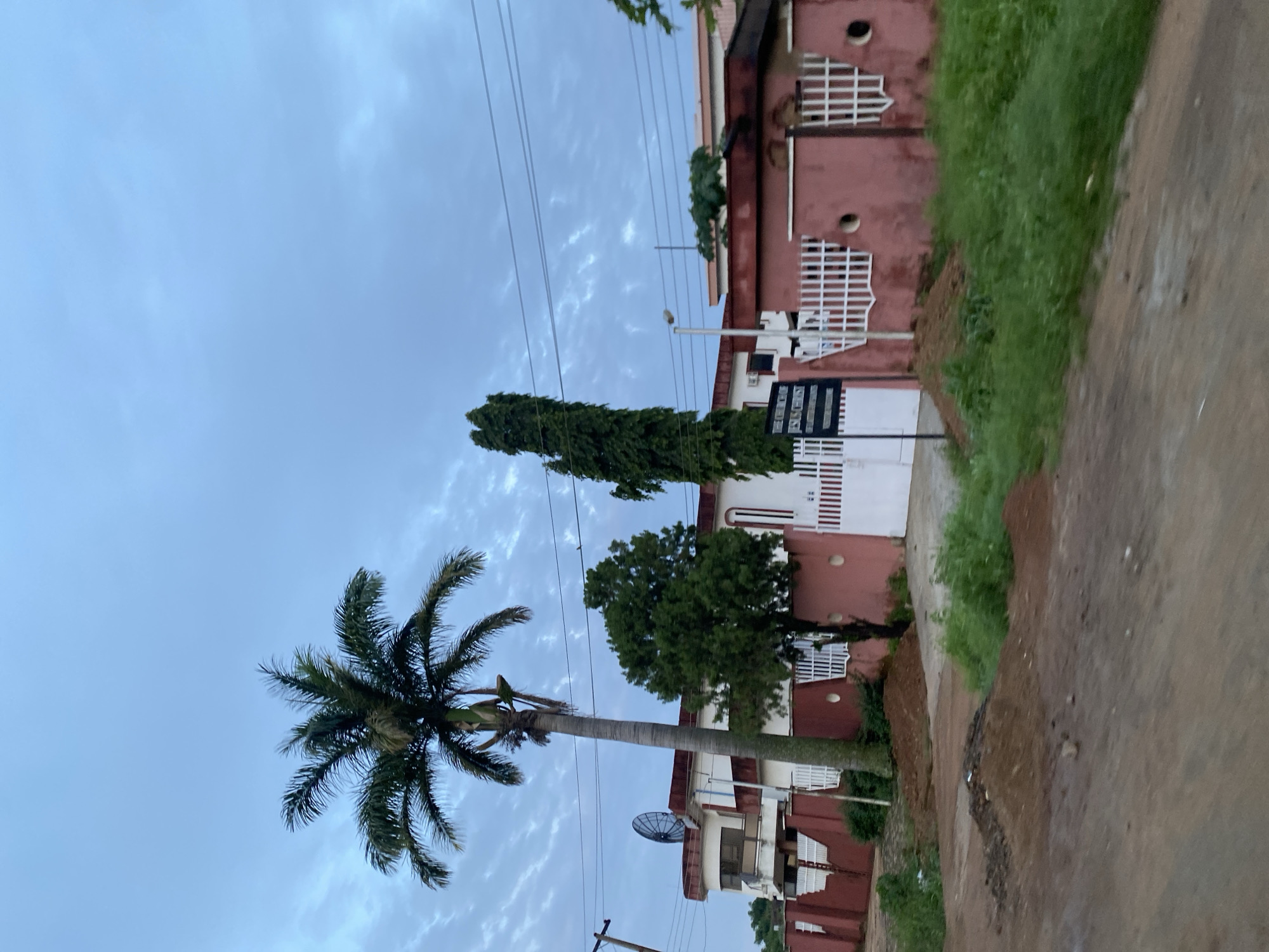 The image depicts a street view in Jos, Nigeria, on a cloudy day. In the foreground, a dirt road with piles of brown soil suggests recent activity or natural erosion. To the left, a utility pole with wires stands tall. A red and white building with a curved facade is visible behind a red wall topped with white decorative elements. A large satellite dish is mounted on the roof of this building. A tall palm tree with green fronds dominates the left side of the frame. Adjacent to the building, a white gate is flanked by the red wall. Further to the right, another building, also with red and white accents, is partially visible, featuring a white picket fence. A slender, tall tree with dense green foliage stands between the two buildings. A signpost reads "THE CHURCH OF JESUS CHRIST" with additional text that is not legible. Green grass and vegetation grow along the roadside and in front of the walls. The sky is overcast with scattered clouds, casting a muted light on the scene. The overall impression is that of a residential or institutional area in an urban setting.