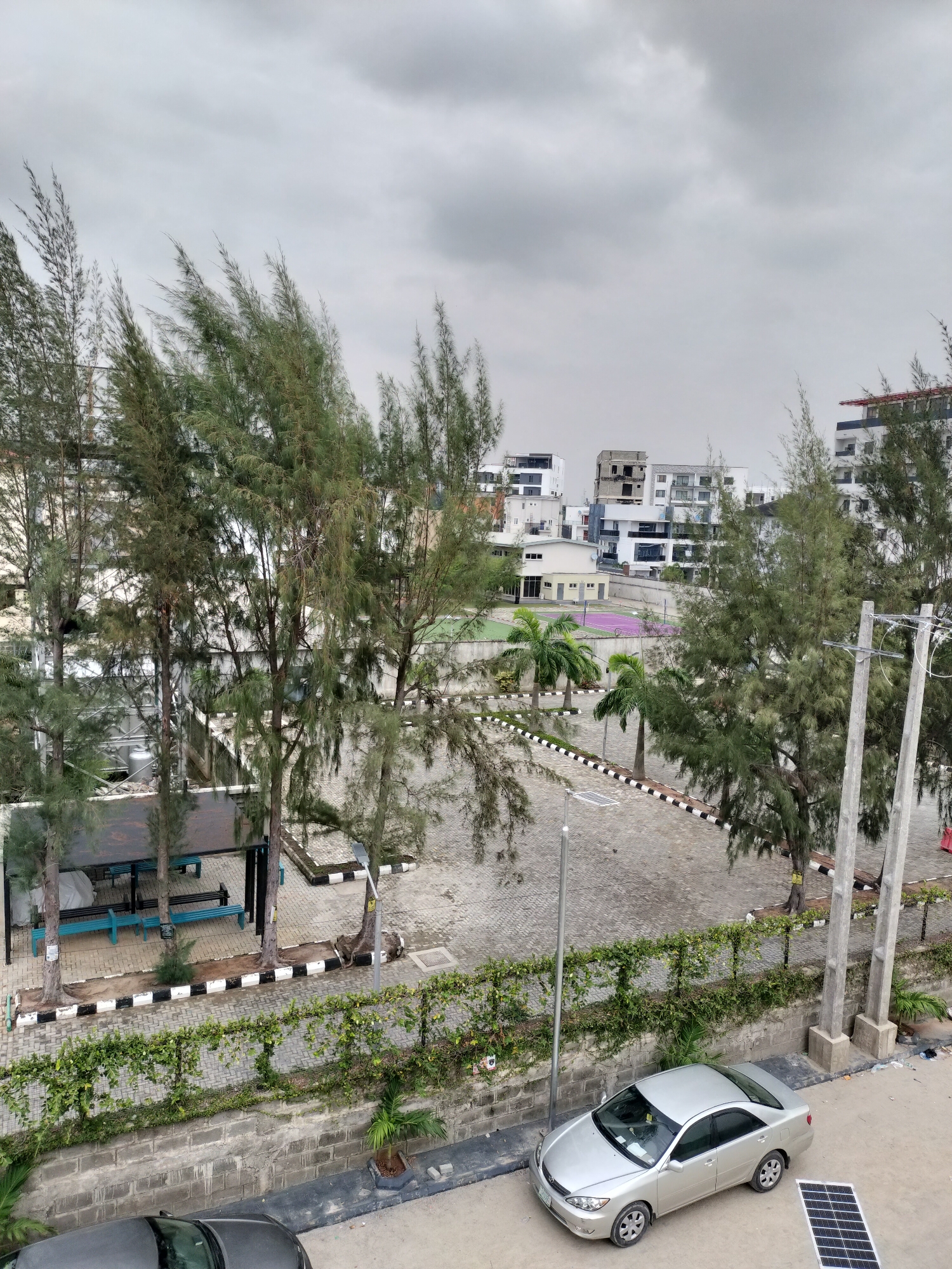 The image captures an elevated view of an urban scene in Moba, Nigeria on an overcast day. In the foreground, a silver Toyota Camry is parked near a gray brick wall covered with green vines. A black solar panel lies on the ground near the car. A covered seating area with teal benches is visible further back, surrounded by tall, slender trees. In the background, modern white buildings and a purple tennis court can be seen. The street is paved with interlocking blocks and bordered by black and white painted curbs. Overhead electrical lines are supported by concrete poles on the right. The sky is uniformly gray, suggesting cloud cover.