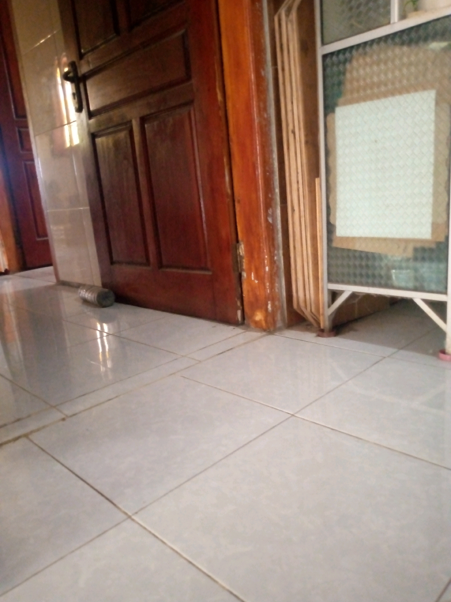 The image depicts a close-up, low-angle view of a tiled floor in front of a dark wooden door in Sedati, Indonesia. The door, with its visible handle and paneling, is slightly ajar. To the right of the door, a decorative screen with a patterned insert is visible. In the foreground, a cylindrical object, possibly a roll of tape or a sealant, lies on the light-colored floor tiles. The overall lighting suggests an indoor environment, possibly a hallway or entrance.