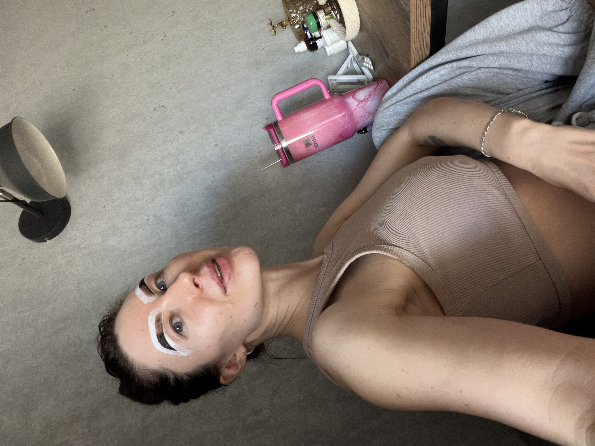 The image shows a woman with tape on her eyebrows, presumably for an eyebrow treatment. She is lying on a grey floor near a table with some small decorative items and a pink Stanley cup. A grey lamp is also visible. There's nothing in the image that suggests it was taken in Perm, Russia.