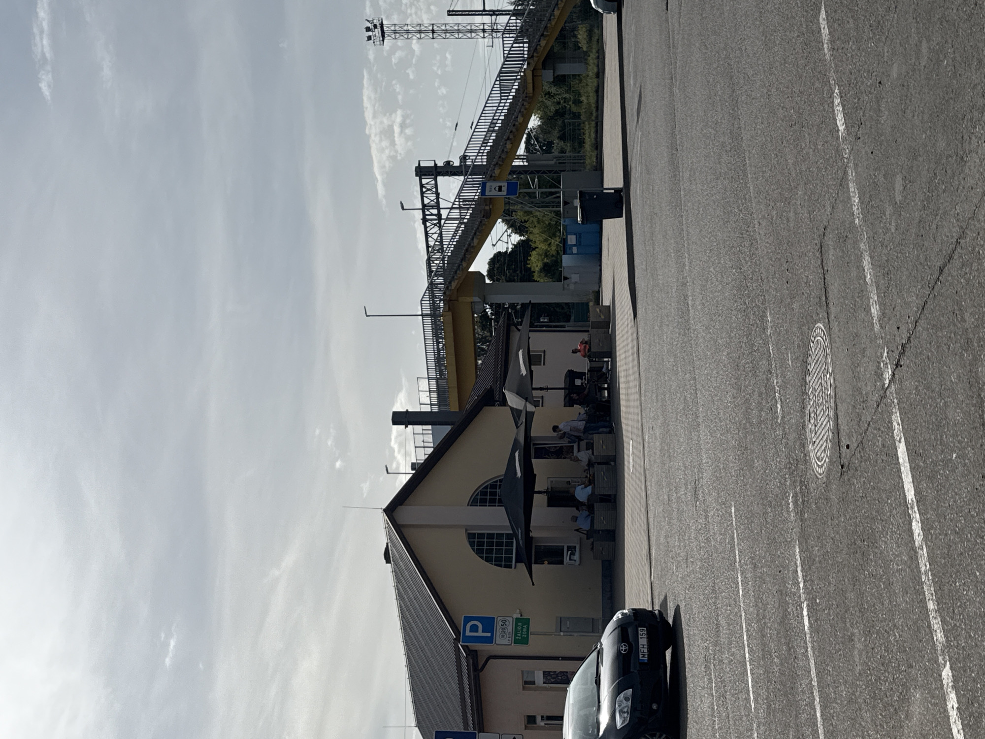 The image shows a street-level view of a building with a yellow facade and a dark-tiled roof, located in Šiauliai, Lithuania. Adjacent to the building is a yellow pedestrian overpass that crosses over railway tracks. Several people are seated at tables outside the building, under black umbrellas. A black car is parked in front of the building. In the foreground, there is a paved road with white lane markings and a manhole cover. The sky is overcast with some clouds.