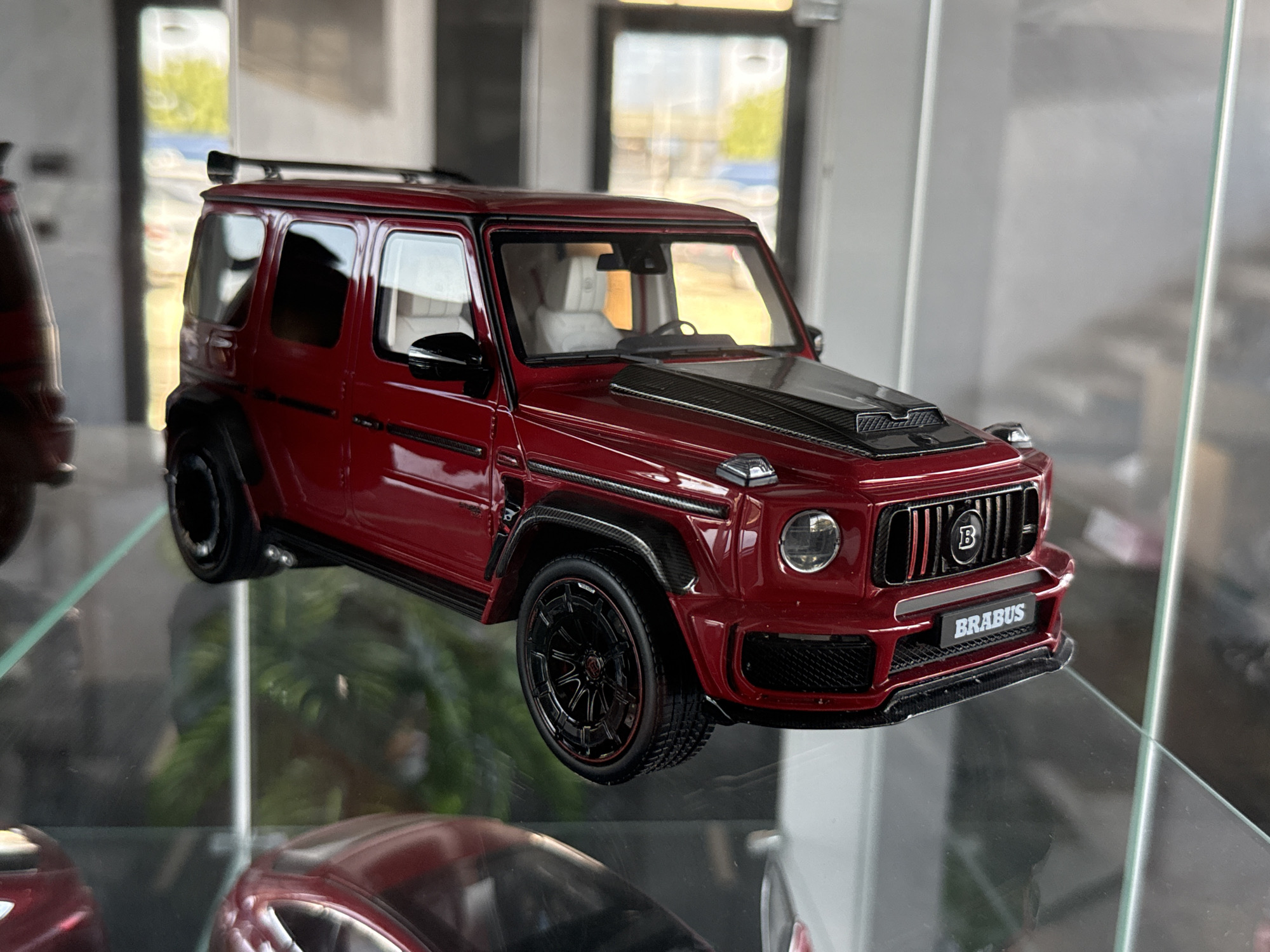 A close-up, eye-level shot shows a detailed, red Brabus G-Wagon model car displayed on a glass shelf. The model has a black hood with carbon fiber accents, black wheels with red rims, and a black roof rack. The interior is visible with light-colored seats. The car is positioned on the right side of the frame, angled slightly towards the viewer. The background is blurred, with hints of daylight and architectural elements, possibly suggesting an indoor display or a showroom. The overall scene is well-lit, emphasizing the glossy finish of the model. There is no discernible location in Plovdiv, Bulgaria mentioned or implied within the image itself.