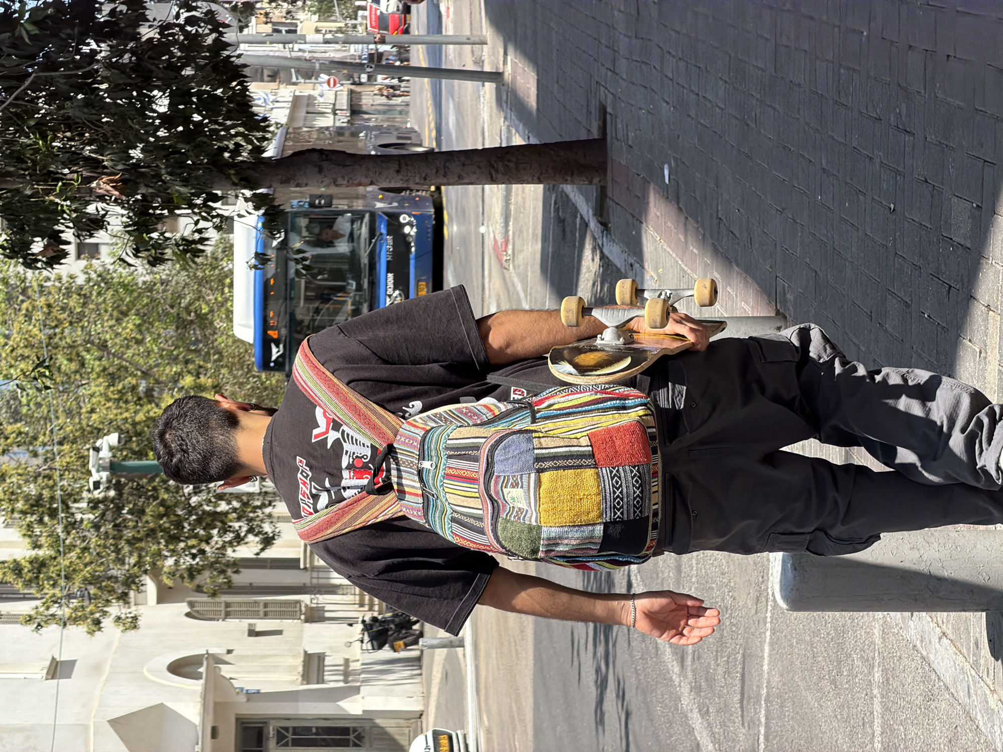 A person stands on a sidewalk in Tel Aviv, Israel, holding a skateboard. The person wears a black t-shirt, grey cargo pants, and a colorful patchwork backpack. A bus is visible on the street behind them. The setting appears to be a sunny urban environment.