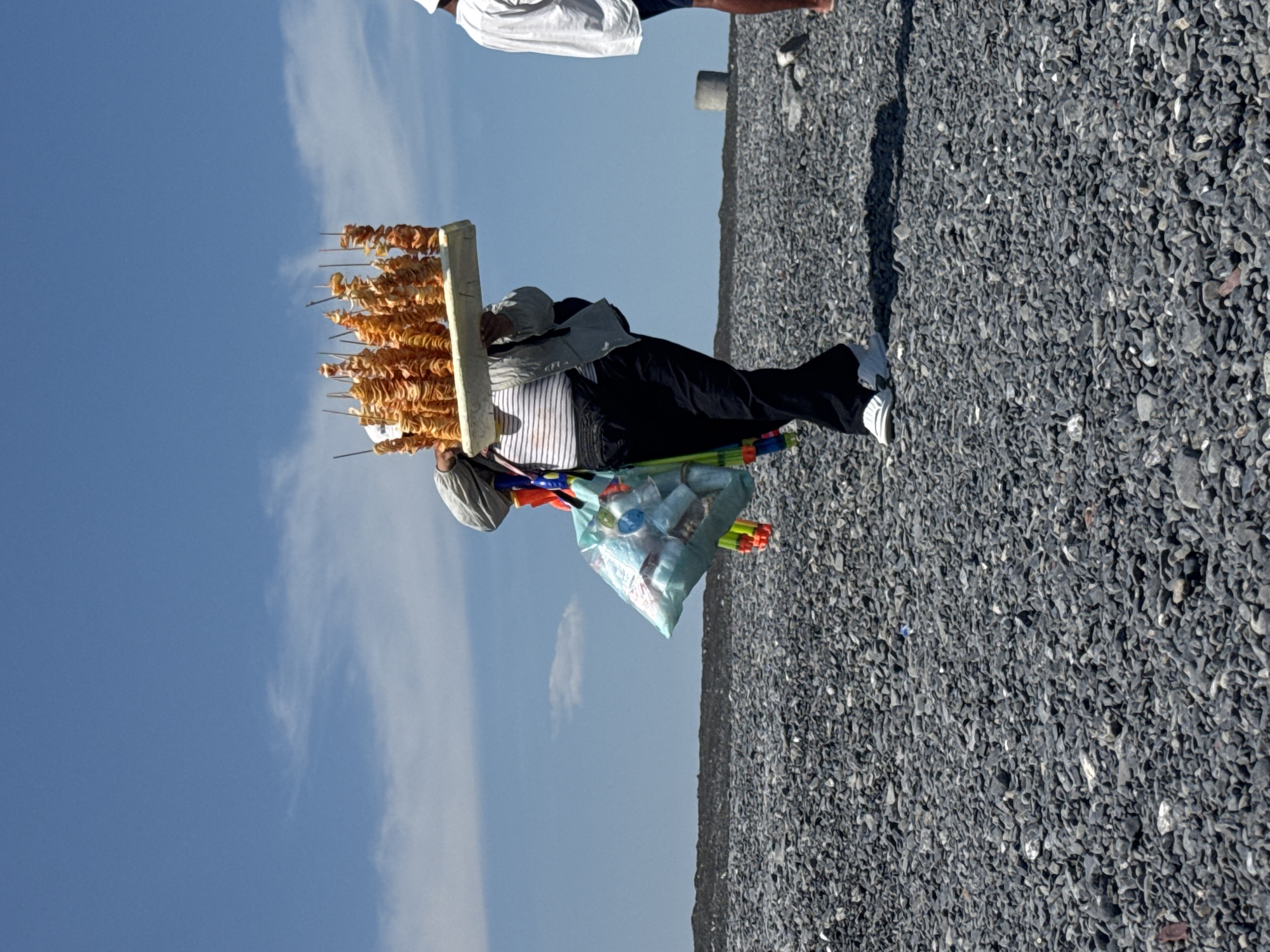 A person is walking on a gravelly surface under a bright blue sky with scattered clouds. They are carrying a tray of spiral-cut fried potatoes on sticks. The person is wearing a striped shirt, a light-colored jacket, black pants, and white sneakers. A bag with colorful items, possibly toys or other merchandise, hangs from their shoulder. Another person in a white shirt and dark shorts is visible to the right. The environment appears to be an outdoor, possibly coastal or industrial area, with a dark line of landscape in the background. There are no discernible buildings or specific landmarks to pinpoint a location within the city of Ақши, Kazakhstan, though the scene suggests an open, perhaps somewhat barren, area.
