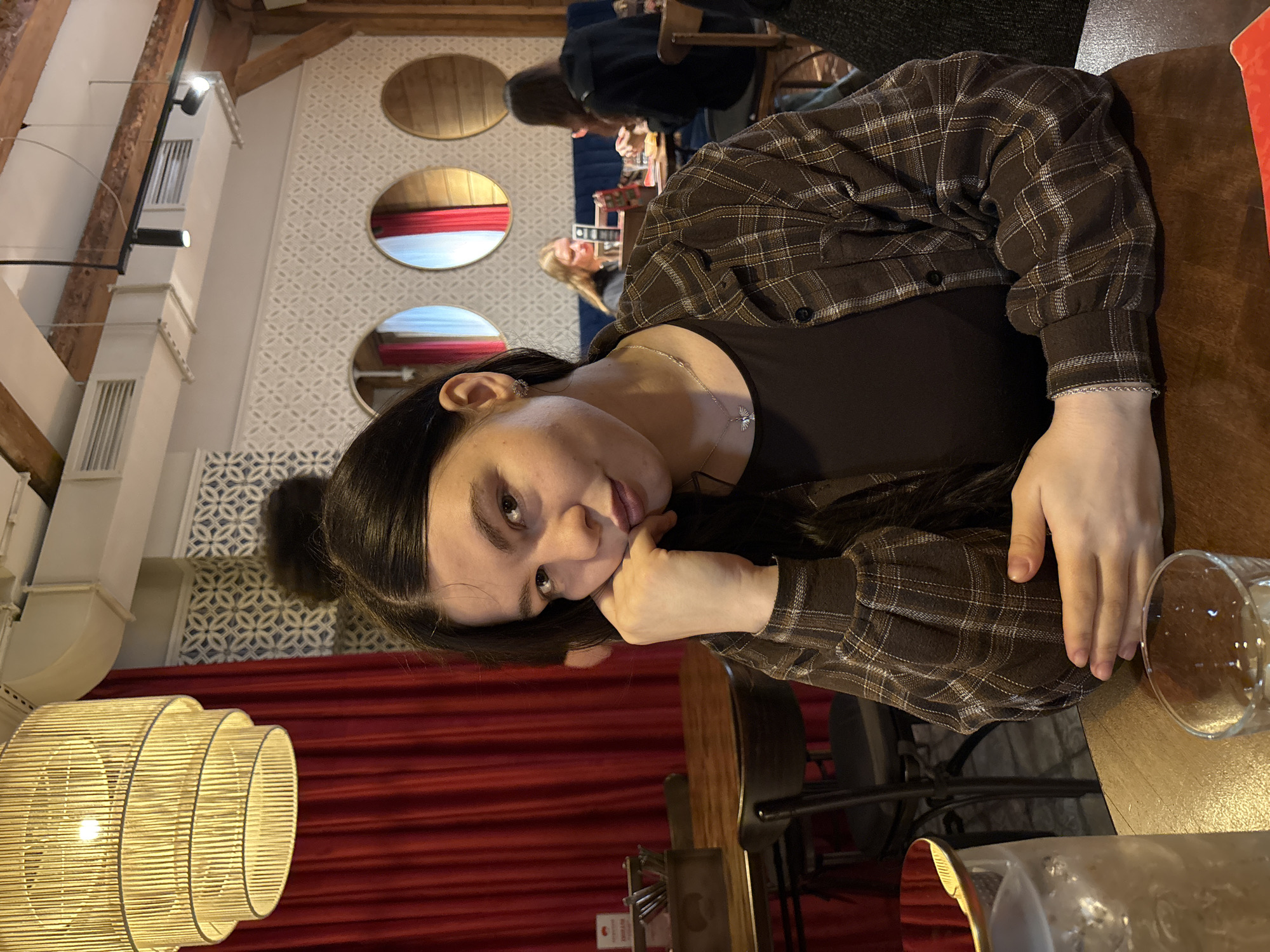 The image shows a woman sitting at a table in a restaurant or cafe. She is wearing a brown plaid shirt over a dark top, and a silver necklace is visible. She is resting her chin on her hand and looking at the camera. A glass is on the table in front of her. In the background, other patrons are seated at tables, and there are decorative circular windows on the wall. A large, woven light fixture is visible in the foreground. A red curtain is also present. The location may be in the city of Perm, Russia.