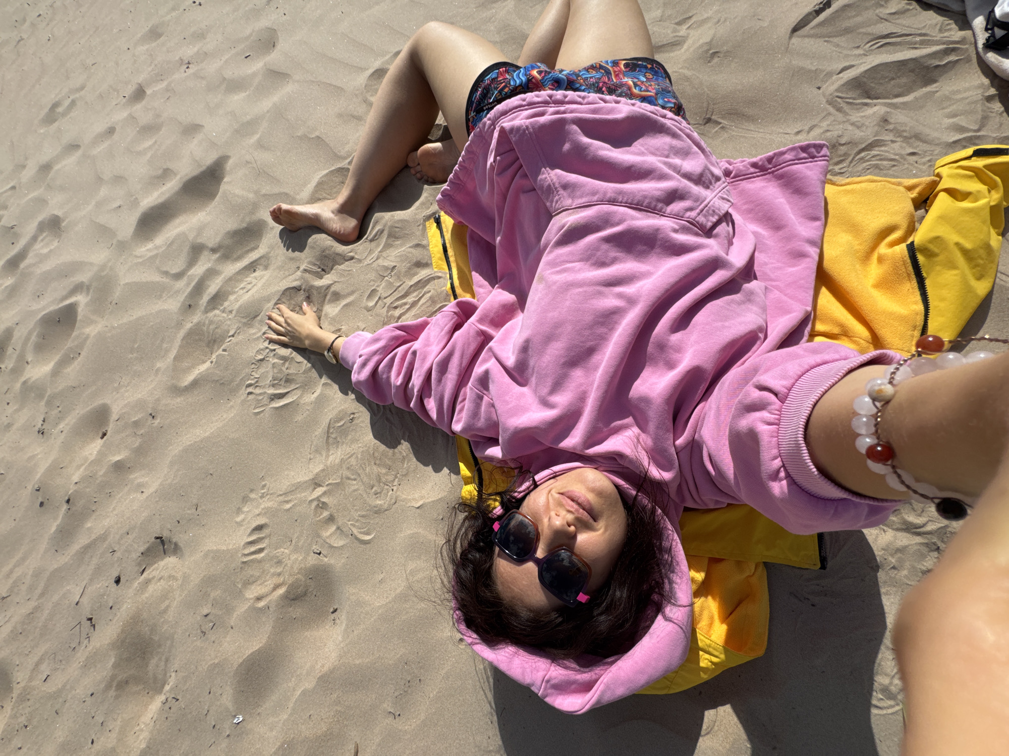 A woman is lying on a sandy beach, on top of a yellow towel and a pink hoodie. She is wearing sunglasses and colorful shorts, and has a beaded bracelet on her wrist. The woman is holding a camera to take a selfie. The sun is casting shadows on the sand.