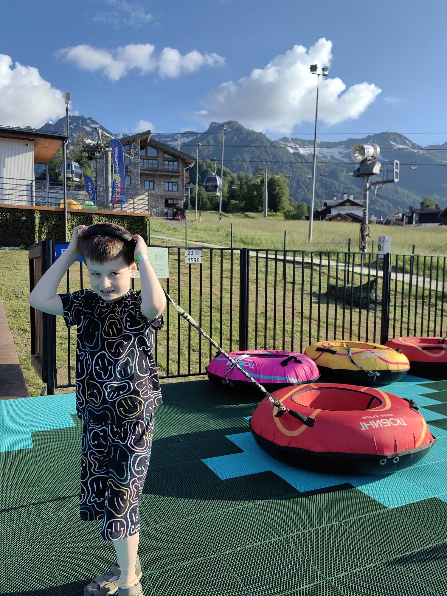 A young boy stands on a colorful mat, his hands on top of his head, looking at the camera with a slight smile. He is wearing a black t-shirt and shorts with a pattern of various smiley faces in white, yellow, and pink. The mat he is standing on is green with a blue grid pattern beneath it. In the background, several inflatable tubes in red, yellow, and pink are arranged on the mat. A black metal fence separates the mat area from a grassy field. Beyond the fence, a building with a stone facade and large windows is visible, with mountains and ski lifts in the distance. The sky is blue with scattered white clouds, suggesting a clear and sunny day. A sign with Cyrillic text and numbers is visible on the fence. The overall scene appears to be an outdoor recreational area, possibly at a resort, given the mountains and ski lift infrastructure. The time of day is likely afternoon due to the sunlight and shadows.