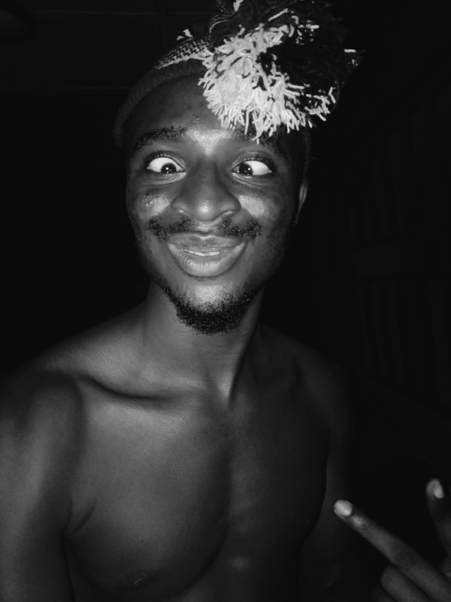The image is a black and white portrait of a young Black man, captured in a playful and humorous moment against a dark, indistinct background. He is shirtless, revealing his muscular chest and shoulders. He wears a beanie-style hat adorned with a large, fluffy, textured pom-pom, prominently positioned on the left side of his head, with some strands cascading over his forehead.
The man's expression is comical: his eyes are crossed, looking inward, and his lips are puckered into an exaggerated, wide smile. He has a neatly trimmed beard and mustache. His skin, particularly his forehead, shows some texture or marks, possibly sweat or smudges, illuminated by strong, direct lighting.
In the lower right corner, part of his right hand is visible, making a peace sign (V-sign) with his index and middle fingers extended. The lighting appears to be artificial and directly focused on the subject, creating highlights on his skin, eyes, and the pom-pom of his hat, while plunging the background into deep shadow. The overall mood is lighthearted and jovial. There is no visible text in the image. The location context provided is Onitsha, Nigeria.