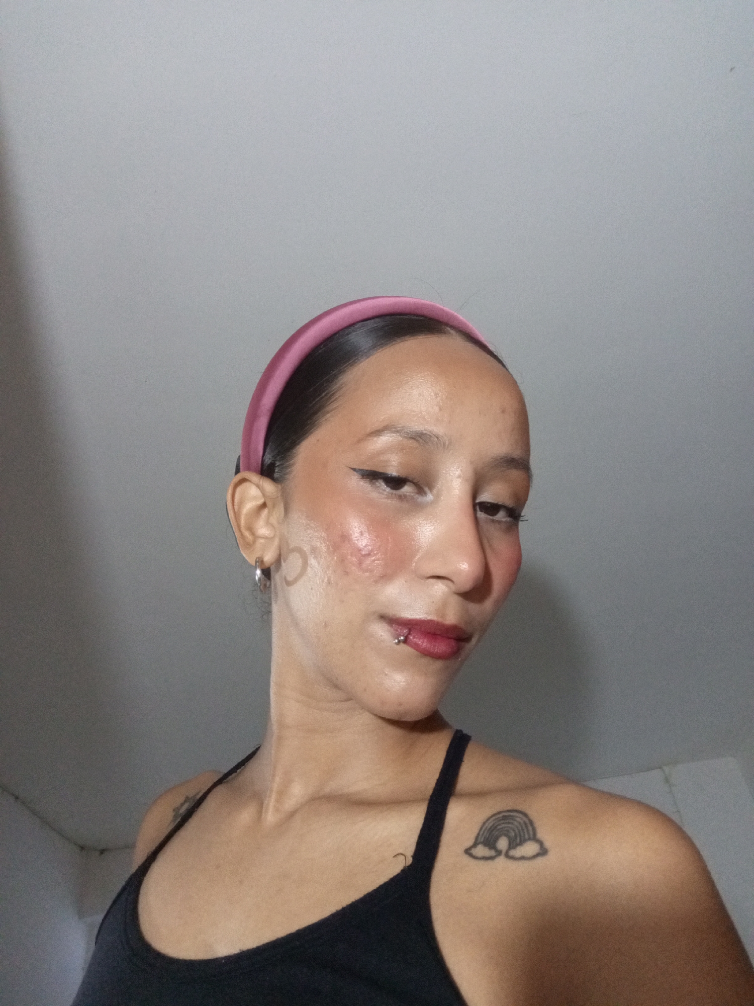 A young woman with tan skin, a pink headband, and a black tank top is depicted in a close-up, angled shot. She has dark hair pulled back, noticeable makeup including eyeliner and lipstick, and a piercing on her lower lip. A rainbow tattoo with clouds is visible on her left shoulder. The background consists of a plain, light-colored wall. There is no information available in the image or prompts about the location being Porlamar, Venezuela, or any indicators of a specific city.