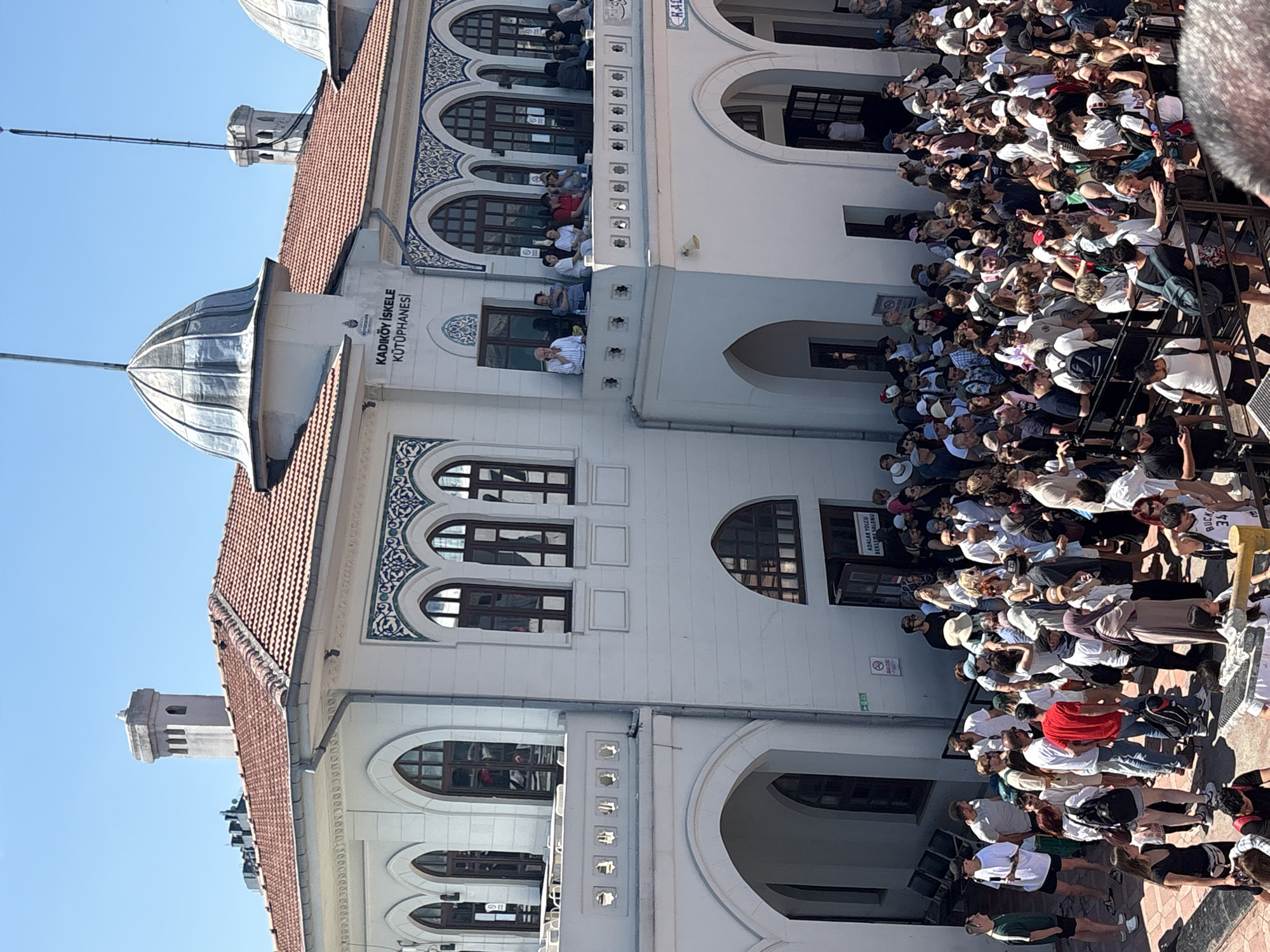 The image captures the exterior of the Kadıköy İskele Train Station and Library in Kadıköy, Istanbul, Turkey, on a sunny day. A large crowd of people, appearing to be tourists and commuters, are gathered outside the station. Many are standing in queues, while others are moving around. The building itself is white with a red tile roof, featuring arched windows and decorative blue tilework. A balcony on the upper level is occupied by a smaller group of people. The scene suggests a busy transportation hub or a popular public space.