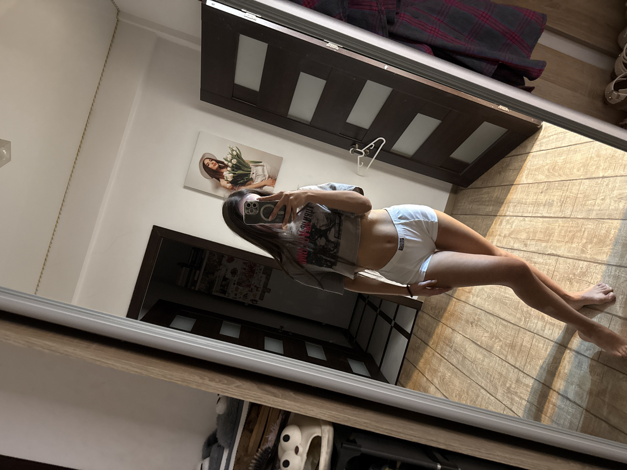 A young woman is taking a selfie in a room. She is wearing a grey cropped t-shirt and white shorts. She is standing in front of a large mirror that reflects her and the room. The room has wooden flooring and a dark brown door with frosted glass panels. On the wall, there is a framed picture of a woman with flowers. To the left of the mirror, there is a shelf with some items, including a pair of novelty slippers. The image was taken in Chișinău, Moldova.
