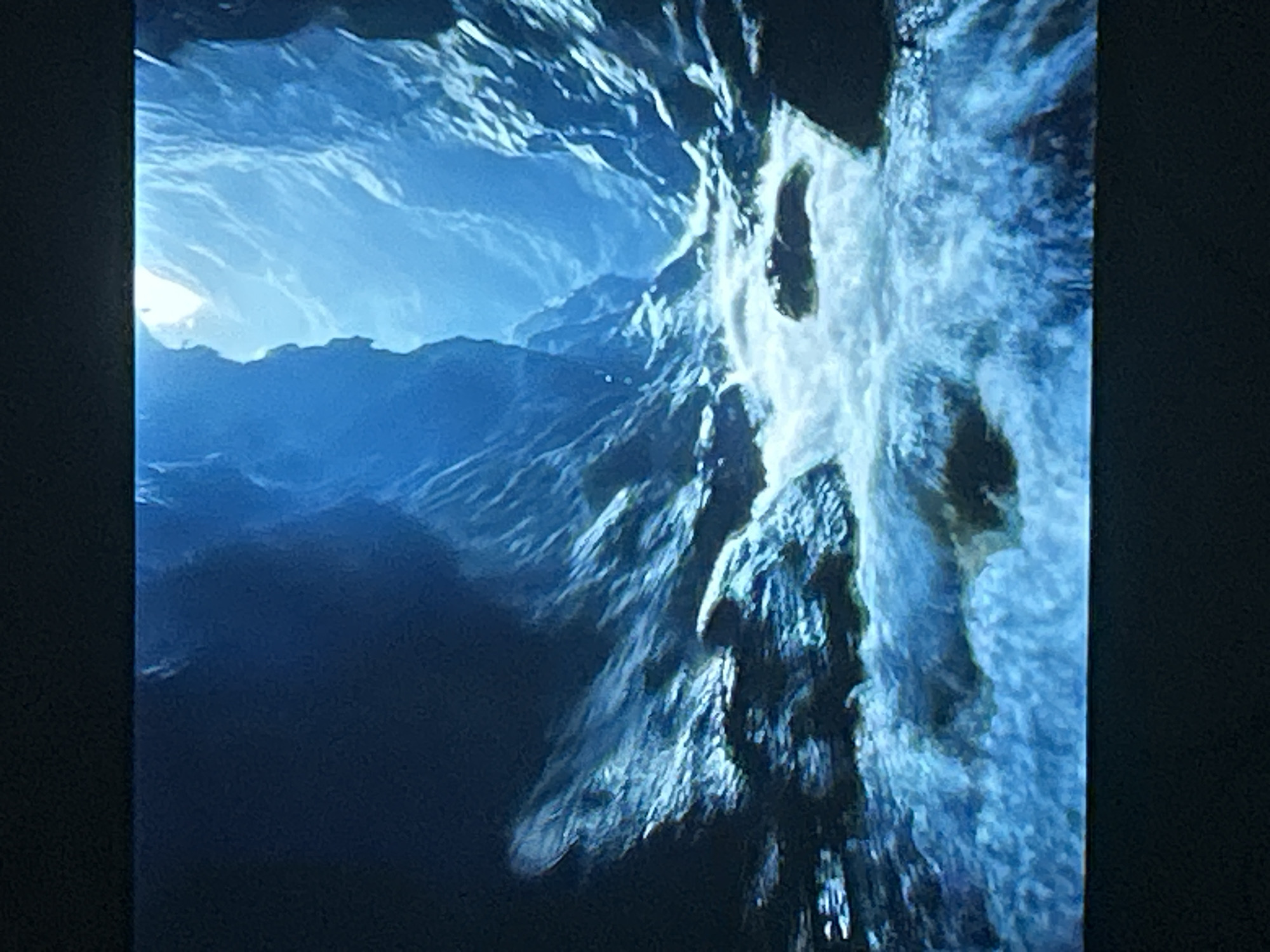 An eye-level medium shot captures a vibrant blue and white digital rendering of a narrow, steep canyon. The canyon walls, textured and jagged, rise dramatically on either side, their surfaces catching the light that streams in from an opening high above. The opening at the top of the canyon casts a bright, ethereal glow, illuminating the scene with a cool, blueish light. At the bottom of the canyon, a turbulent river flows rapidly over rocks. The water is a dynamic mix of white foam and deep blue, suggesting a strong current. The overall impression is one of a natural, albeit digitally rendered, landscape.