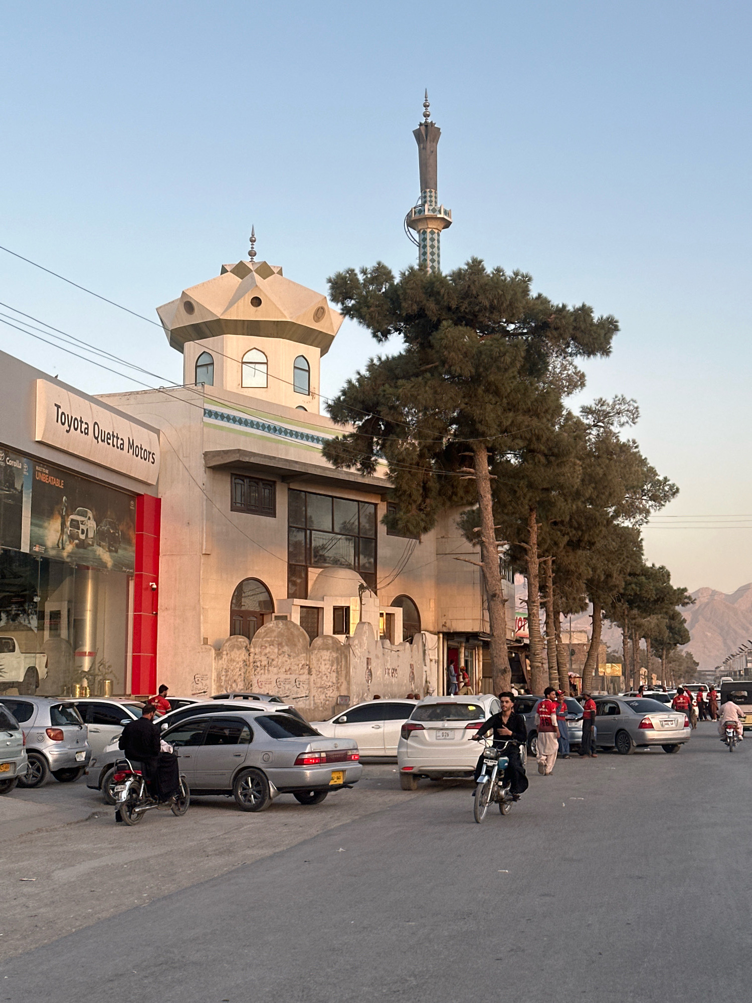 The image captures a street scene in Quetta City Tehsil, Pakistan, likely during the late afternoon or early evening, indicated by the warm, low-angle sunlight casting soft shadows. On the left side of the street, a Toyota Quetta Motors dealership is prominently visible with its signage and a display of vehicles behind glass. Adjacent to the dealership is a building with distinctive Islamic architectural elements, including a minaret topped with a spire and a faceted dome-like structure. The building has arched windows and decorative banding.
Several cars are parked along the street, and a few are in motion. A person is riding a motorcycle, and another is a passenger on a motorcycle behind them. Other individuals are standing and walking on the sidewalk. Tall pine trees line the street, their branches reaching towards the sky. In the background, mountainous terrain is visible, characteristic of the region. The overall atmosphere suggests a busy urban environment with a blend of commercial activity and traditional architecture.