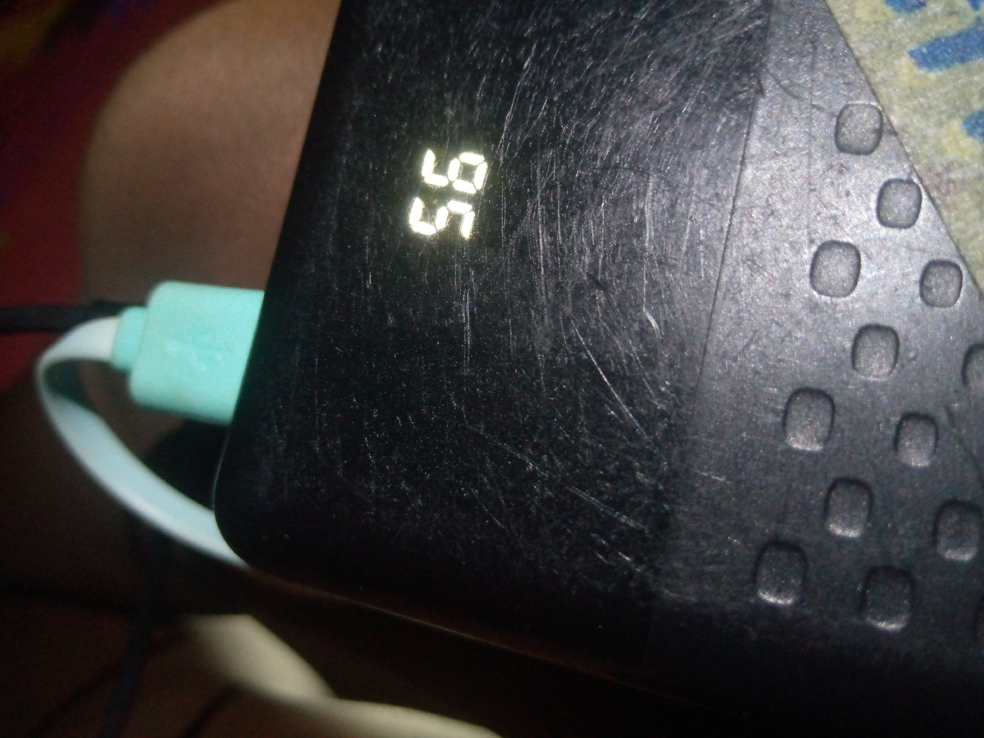 An extreme close-up shot of a black electronic device shows a digital display with the number 56 illuminated in white LED segments. The device's surface is heavily scratched. To the left of the device, a portion of a turquoise USB cable is visible, partially obscured by the edge of the device and a white connector. The background is out of focus and appears to be a mix of brown and red tones, suggesting a domestic or casual setting. There are no people visible in the frame.
