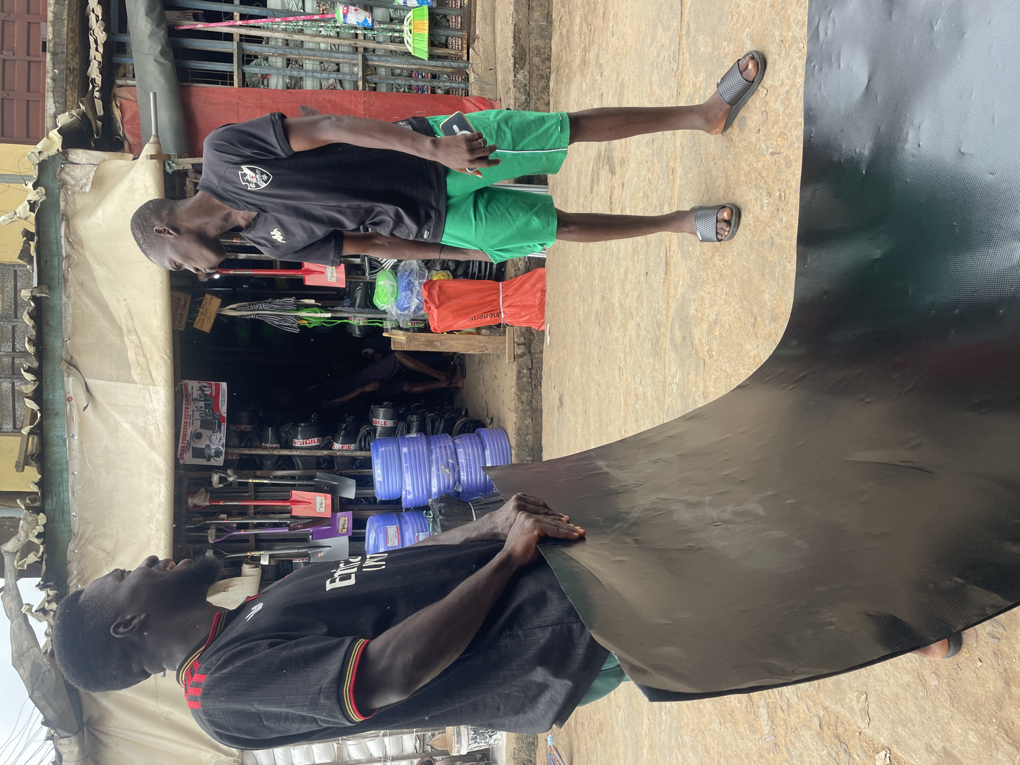 Two men stand outside a storefront in Ogidi, Nigeria, engaged in a discussion while holding a large, black, textured sheet of material. The man on the left, wearing a black shirt with red and yellow trim and a beard, looks towards the right, his mouth open as if speaking. He is holding the left side of the sheet. The man on the right, wearing a black shirt with a white crest and green shorts, looks towards the left, his phone held in his right hand. He is standing near the right side of the sheet, which is spread out on the ground. The background reveals a market stall with various items for sale, including stacks of blue hoses, shovels, and other tools. A sign with text and images is visible on the stall. The ground is paved with what appears to be concrete or stone. The overall scene suggests a commercial interaction or transaction taking place in a bustling outdoor market environment.