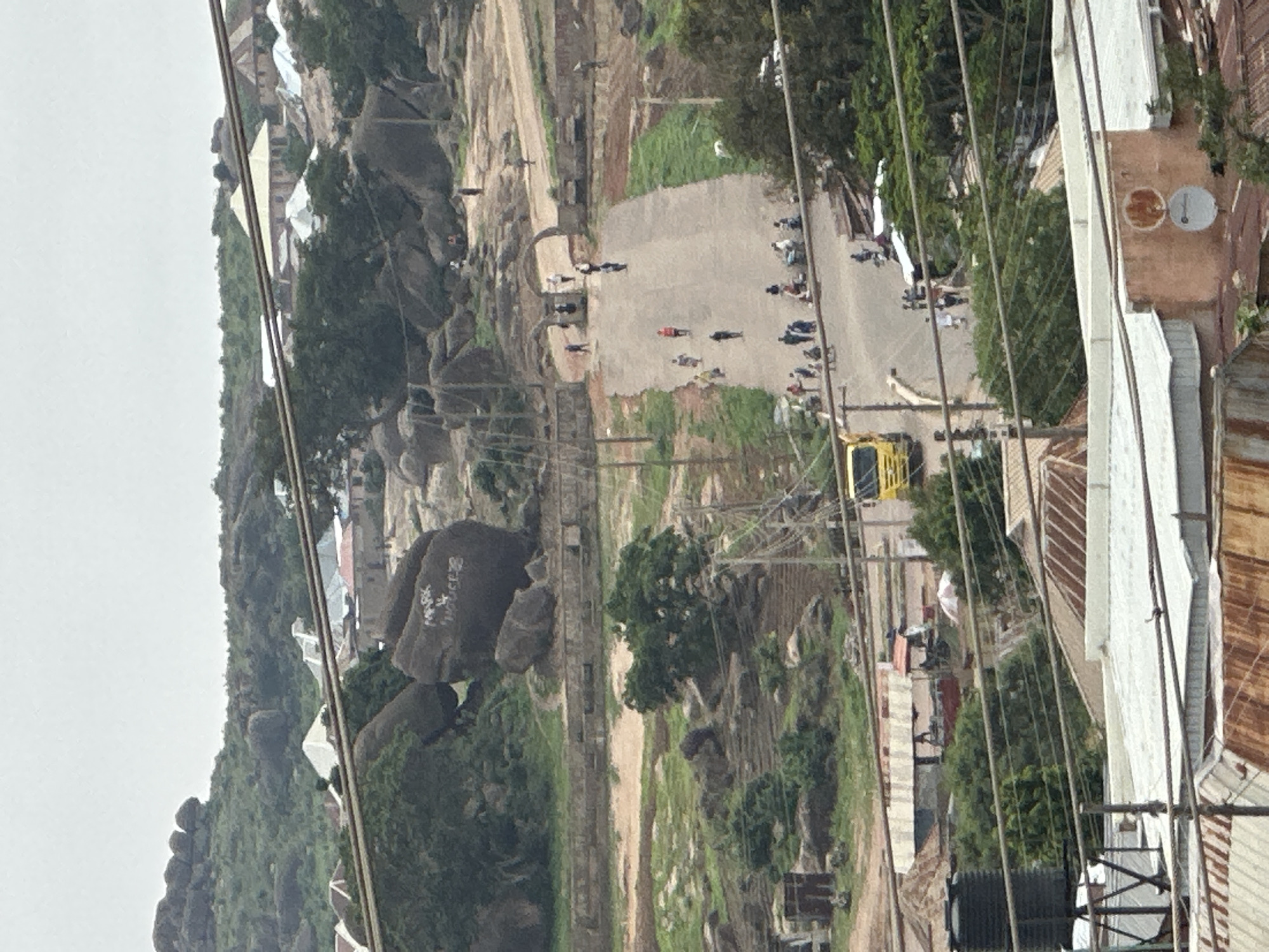 The image depicts a scene in Jos, Nigeria, featuring a rocky, hilly terrain with scattered buildings and a paved road. In the foreground, several rooftops are visible, some with corrugated metal and others with lighter-colored roofing materials. Utility poles and overhead wires crisscross the scene. A large black water tank is situated on the left side, near some structures.
Further into the image, a yellow truck is parked on the side of the road. The road itself winds uphill, leading towards an archway structure. Several people are walking on the road and along the sides, with some appearing to be gathered near stalls or shops. The landscape is characterized by large boulders and green vegetation, including trees and shrubs. In the background, more houses and rocky outcrops are visible, with a hazy sky overhead. Some of the large rocks have graffiti on them. The overall impression is of a bustling, somewhat informal urban environment integrated with a rugged natural landscape.
