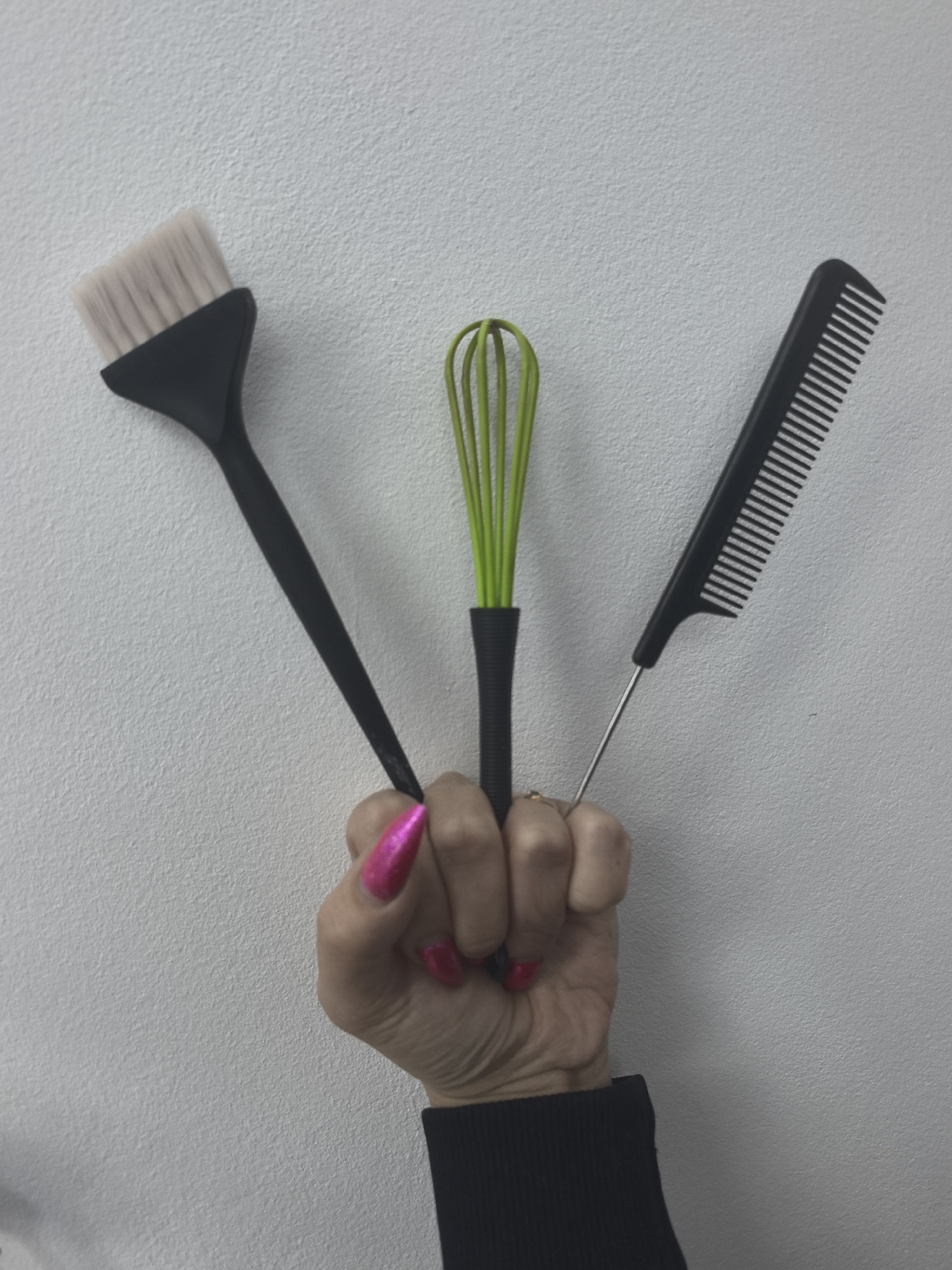 A human hand holds three hair care tools against a plain, light-colored textured wall. The hand, likely female, displays vibrant, reflective pink almond-shaped fingernails, which are notably long on the index and middle fingers. A golden ring is visible on the ring finger, and a black sleeve or cuff is seen at the wrist.
The tools are arranged fanned out from the hand: to the left is a black hair dye brush with soft white bristles. In the center, a lime green mini whisk with a black handle is held. To the right, a black comb with fine teeth and a long, thin metal "rat tail" handle completes the set. These objects are characteristic instruments for hair coloring and styling, suggesting the context of a beauty professional or a personal hair care routine. The whisk is typically used for mixing hair dyes or treatments, the brush for application, and the comb for parting and sectioning hair.
The setting is an indoor space, indicated by the simple, undecorated wall background. No specific time of day, weather, or emotions are discernible. The image captures a practical arrangement of tools for cosmetology, potentially in Chaikovsky, Russia. There is no visible text.