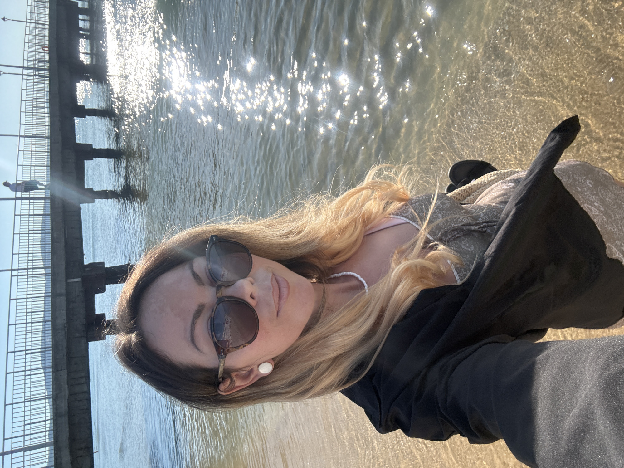 A woman, wearing sunglasses, a black jacket, and a dress, stands on the shore of the sea. The sun glints off the water. A pier with people and lampposts extends into the sea in the background. The location is Nesebar, Bulgaria.