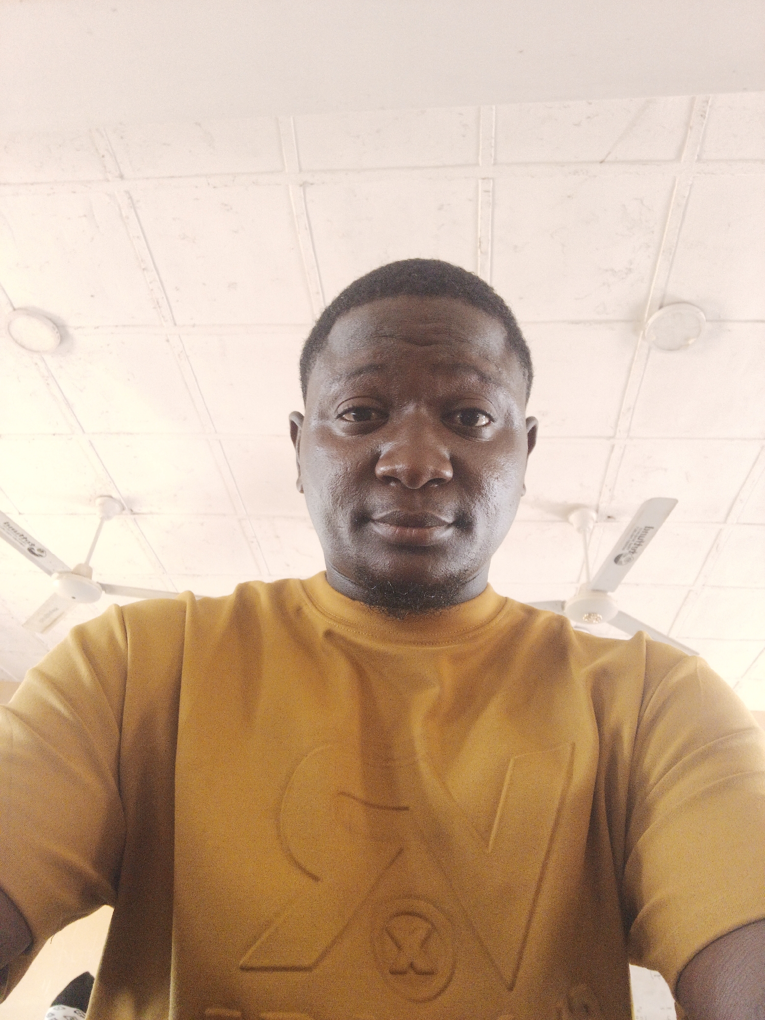 A medium shot of a Black man in a yellow t-shirt. The man is facing forward with his head at the top of the frame, with his shirt in the middle, and a ceiling with ceiling fans visible in the background. The location appears to be an indoor room in Akwanga, Nigeria.