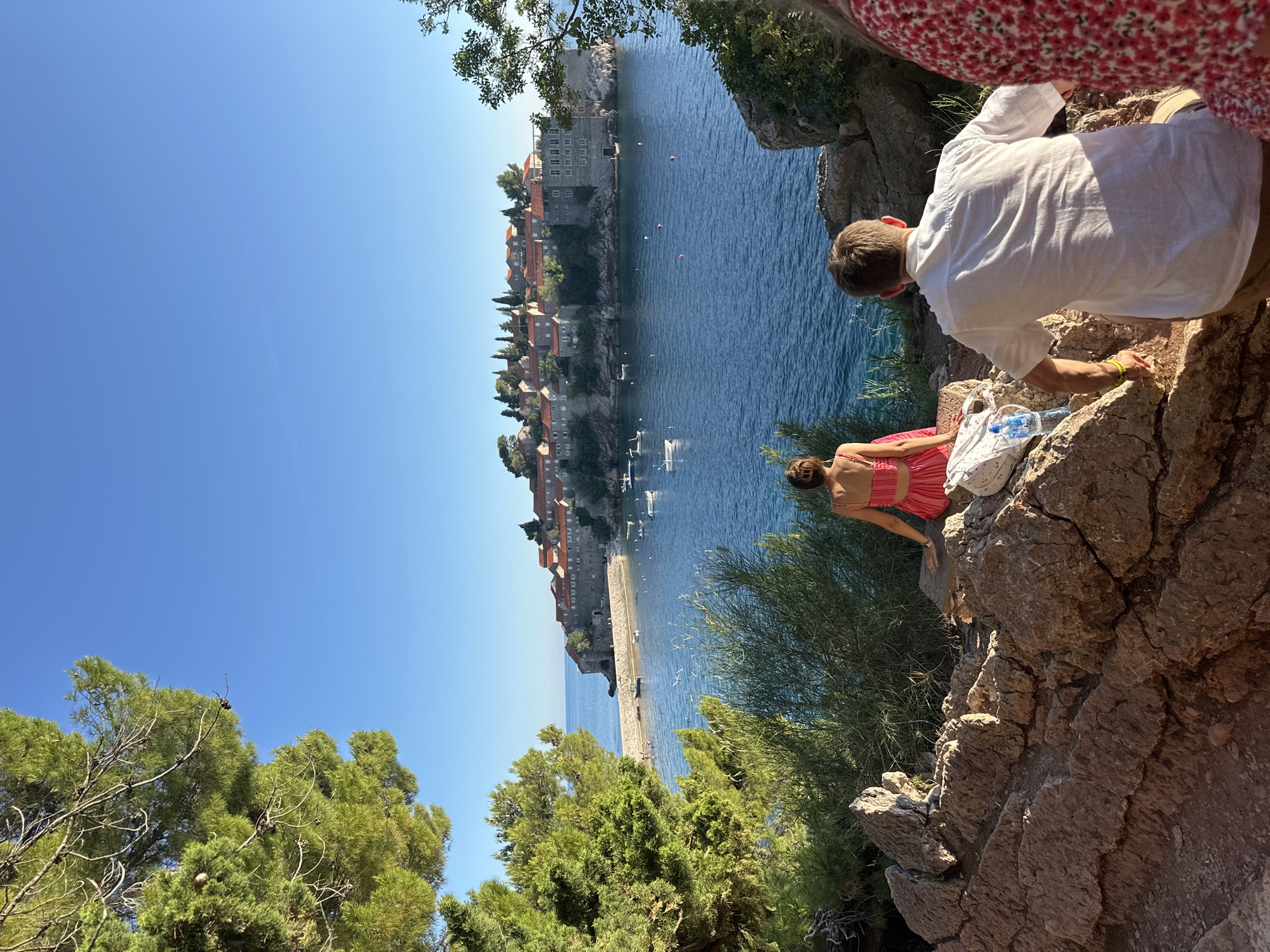 The image shows a scenic view of Sveti Stefan, Montenegro. In the foreground, two people are sitting on a rocky outcrop overlooking the turquoise sea. One person is wearing a white linen shirt and beige shorts, while the other is wearing a pink dress. The person in the pink dress appears to be looking out at the water. The background features the iconic island of Sveti Stefan, with its ancient stone buildings and red-tiled roofs, connected to the mainland by a causeway. The sea is calm with several small boats anchored offshore. Lush green trees and bushes frame the scene. The sky is a clear, bright blue, suggesting a sunny day. The overall mood is relaxed and picturesque, typical of a summer vacation. The time of day appears to be late morning or early afternoon, given the bright sunlight.