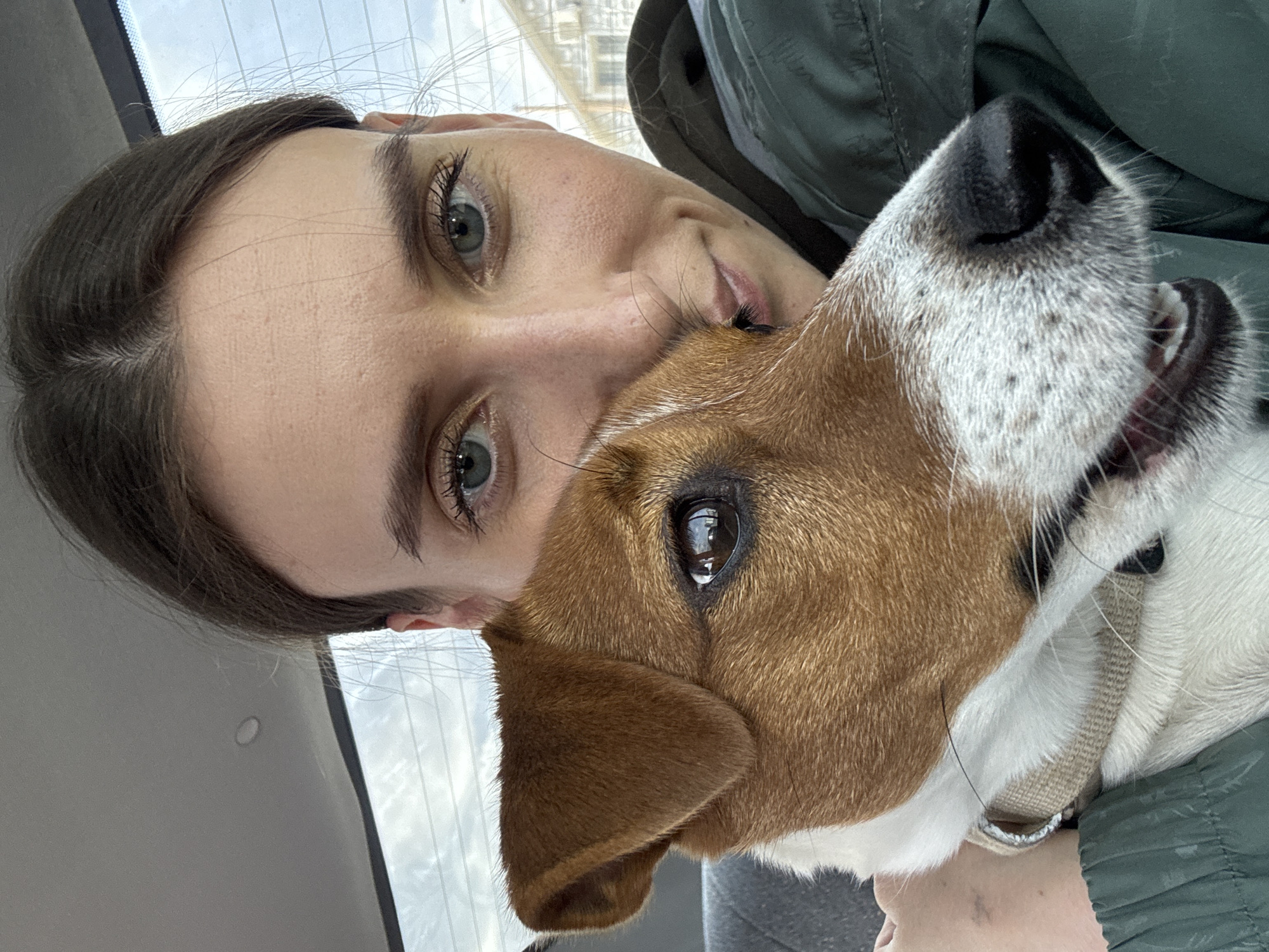 A woman and a Jack Russell Terrier are closely positioned together in what appears to be the interior of a vehicle. The woman has fair skin, brown hair, and green eyes. She is wearing a green jacket. The dog has brown and white fur and is wearing a tan collar. The background shows a blurred urban setting, potentially Chaikovsky, Russia, given the request. A portion of a cloudy sky can be seen through a window.