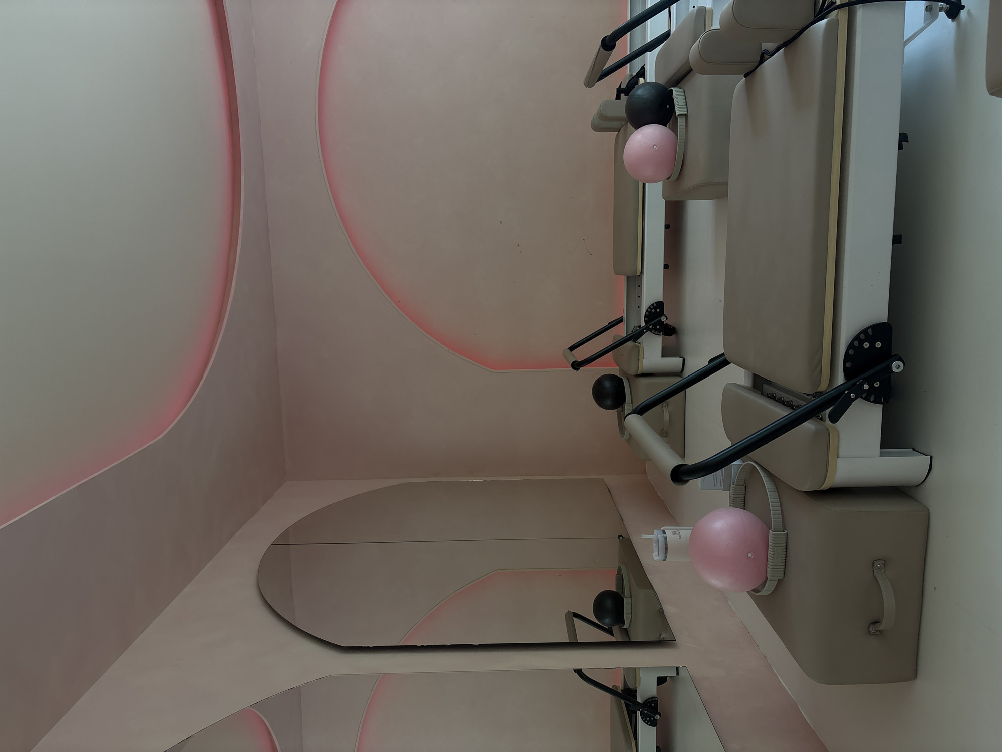 This image shows a pilates studio in Mexico City, Mexico. The studio features several pilates reformers, each with a beige cushioned surface and black metal framing. There are also two large mirrored arches on the left wall, reflecting the reformers and the pink-hued lighting that accentuates the curved ceiling and wall details. On a beige box in the foreground, a pink pilates ball rests next to a grey one. A white water bottle with a straw lid is positioned behind the pink ball. The walls and ceiling are painted in a soft pink color, and the floor is a clean white. The overall aesthetic is modern and serene, suggesting a focus on wellness and exercise.