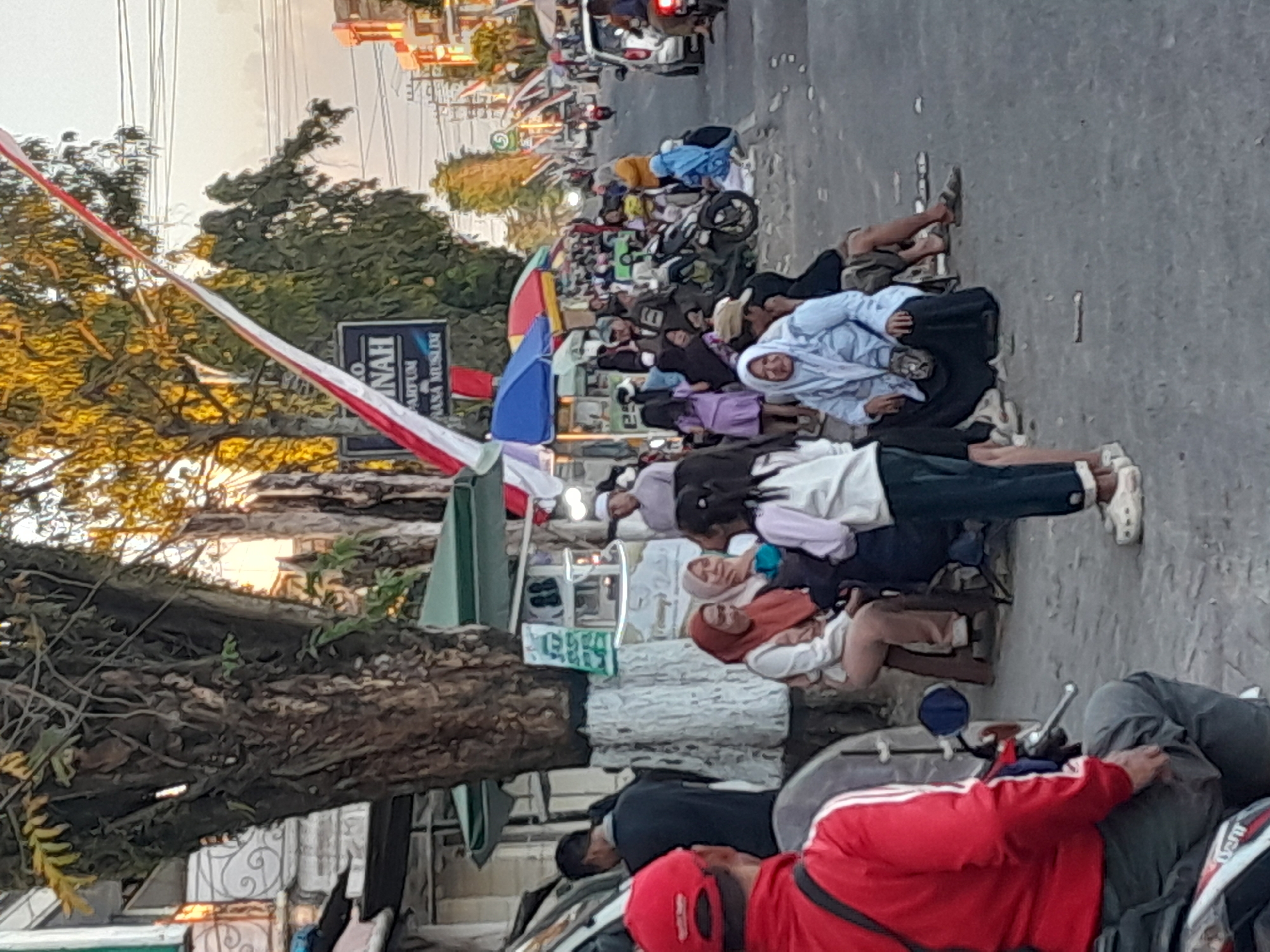 This is a street scene in Gentengwetan, Indonesia, depicting a busy marketplace or gathering area. The foreground features a person wearing a red jacket and a red cap, seated on a motorcycle. To the left, a white van is partially visible.
The middle ground is populated with many people of various ages, mostly dressed in casual attire. Several individuals are seated on stools or directly on the ground. Women are predominantly wearing hijabs, with some in light blue and others in rust-colored headscarves. A young girl in a light-colored jacket and dark pants stands prominently. Tents and stalls are set up along the roadside, suggesting a commercial or community event. Red and white flags, possibly national or celebratory banners, are strung across the scene.
The background shows more people, parked motorcycles, and vehicles, including cars and motorcycles. Buildings line the street, and utility poles with electrical wires are visible. The sky is visible in the upper portion of the image, with a warm, possibly evening light. The overall impression is one of a lively public gathering in a dense urban environment.