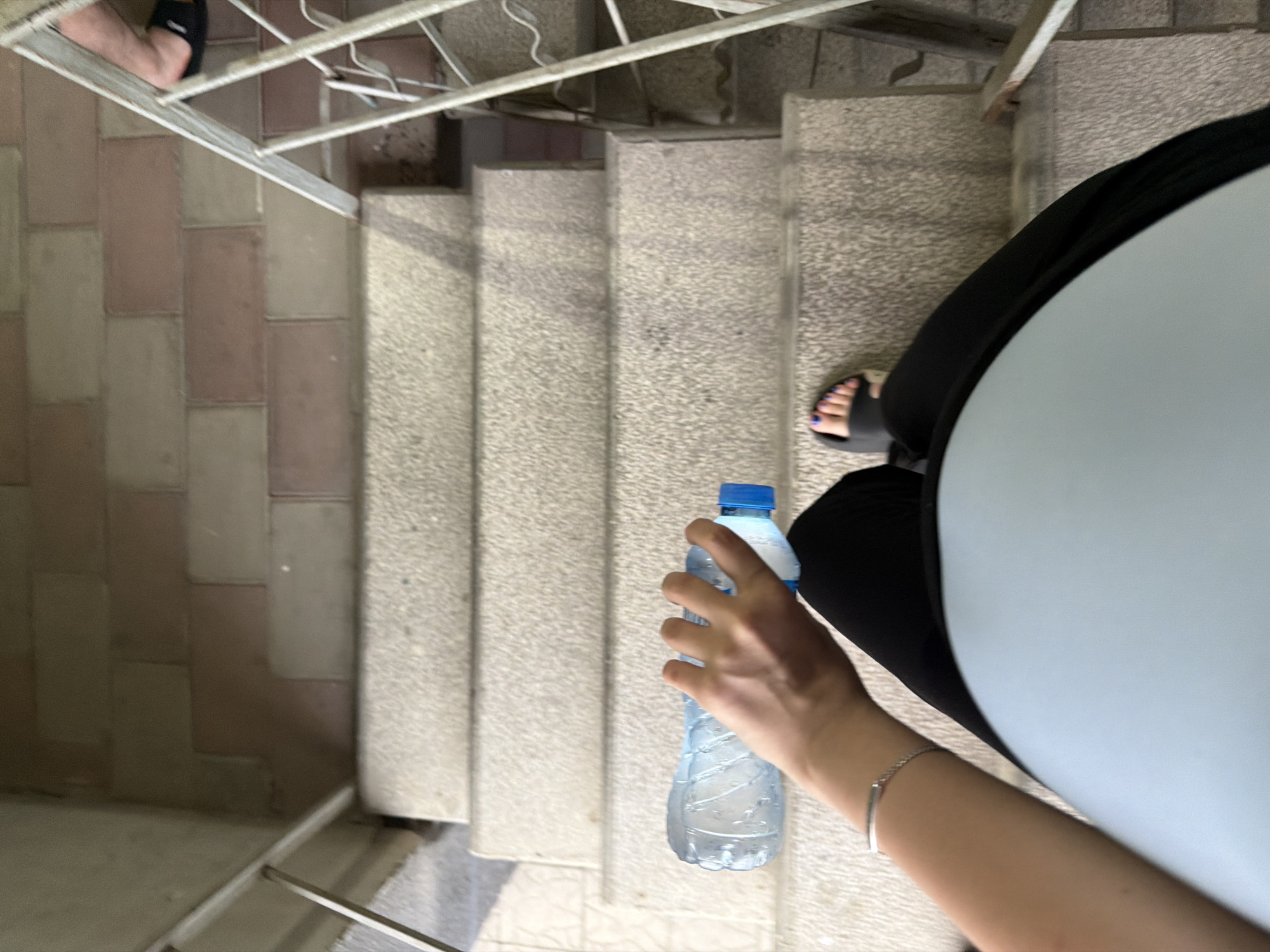 A person is holding a clear plastic water bottle with a blue cap while descending a concrete staircase. The person is wearing black leggings and a light blue top. They are also wearing black slides on their feet, revealing purple nail polish on their toes. A silver bracelet is visible on their wrist. The staircase has a textured, speckled surface and is bordered by a metal railing. To the left of the stairs, the ground is paved with rectangular tiles in shades of pinkish-brown and beige. The overall scene appears to be outdoors, possibly during the daytime given the lighting. The perspective is from above, looking down the stairs. No visible text is present on the water bottle or in the surrounding environment. The setting suggests an urban or residential area with outdoor stair access.