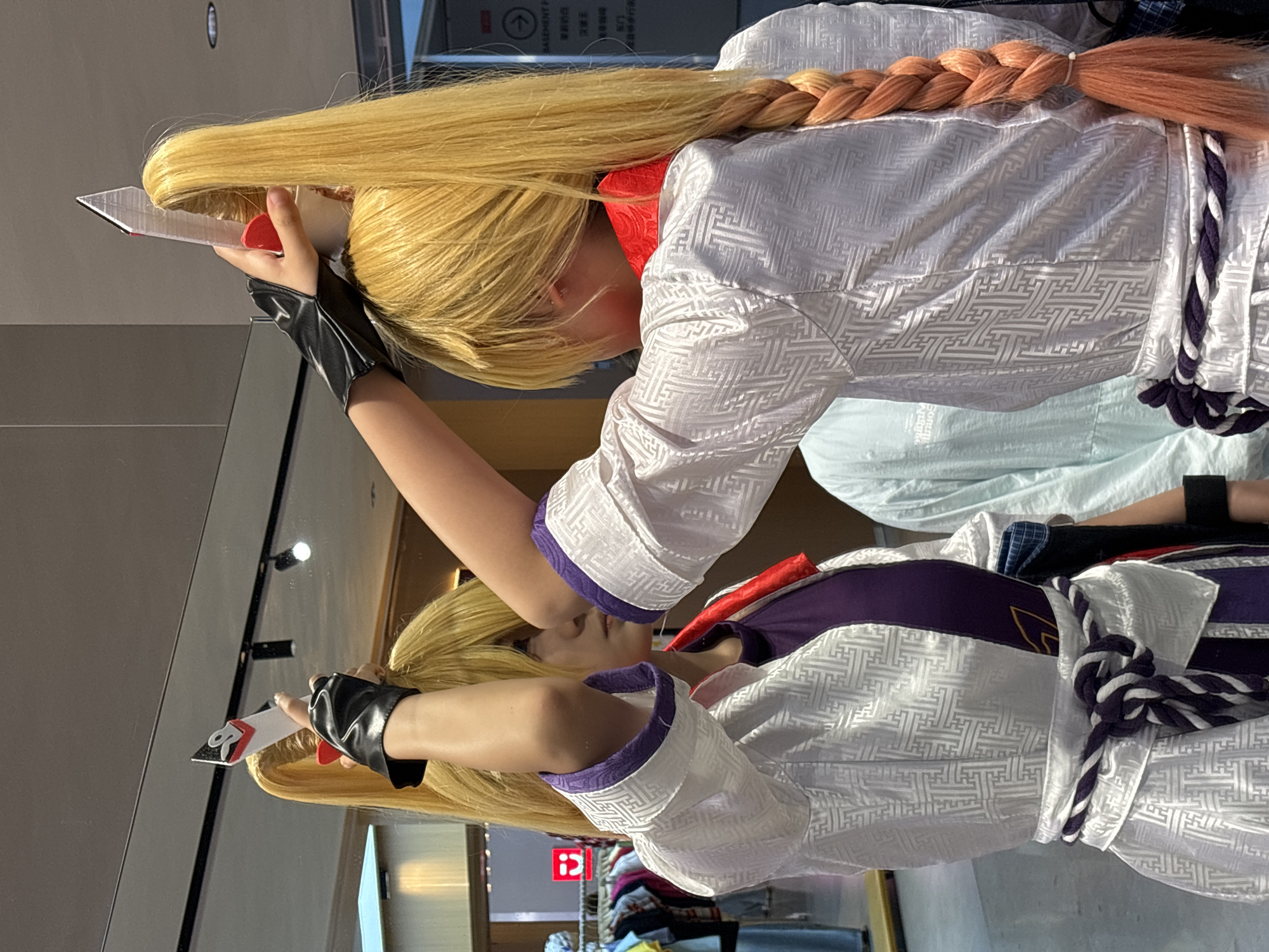 Two people are cosplaying as fictional characters in an indoor location. Both are wearing white and purple patterned tops with black and white rope belts. They both have blonde wigs, one with a braid. They are holding what appear to be prop swords. The environment is a store or shopping area, with clothing racks visible in the background. The specific city and location are unknown.